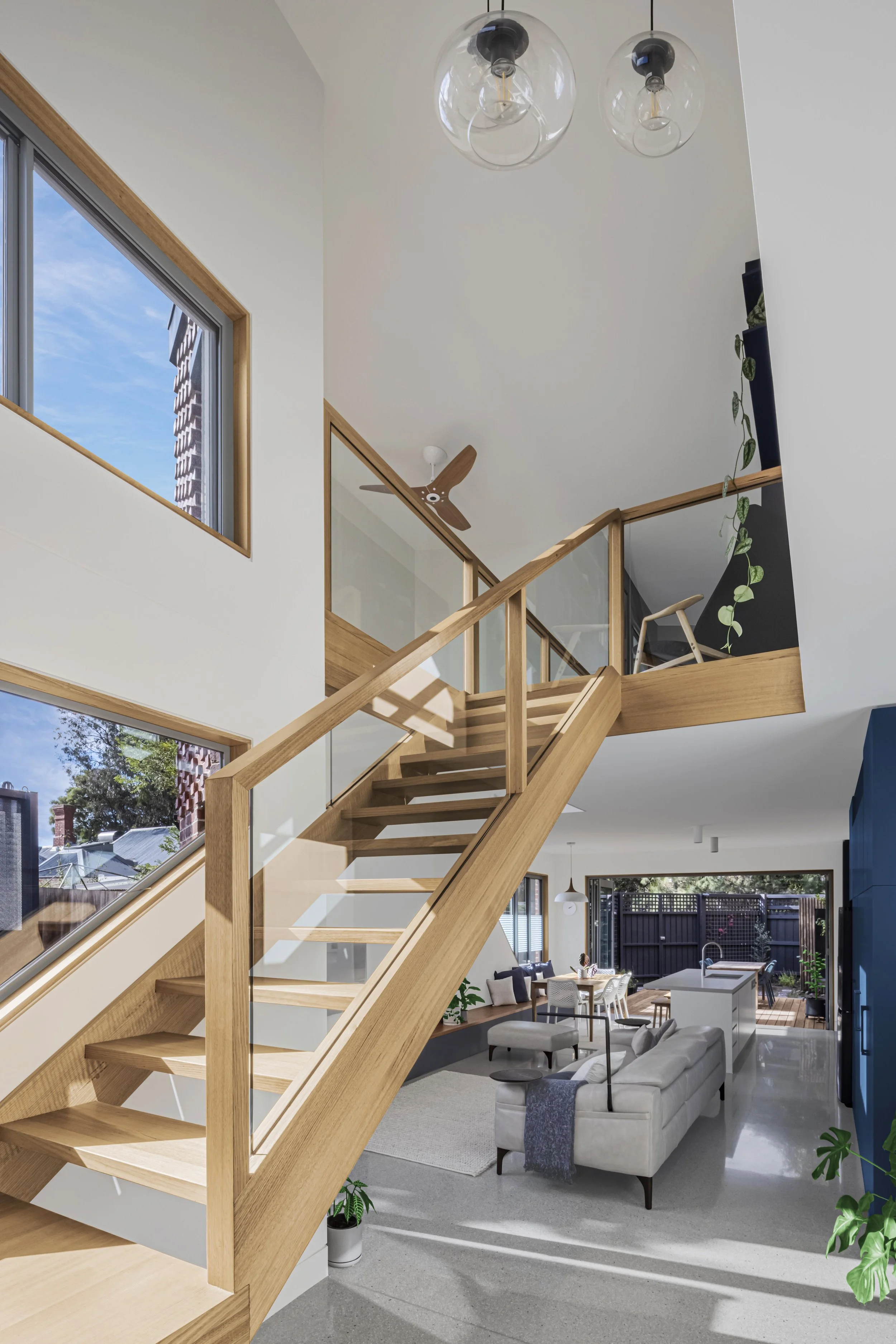 Light-filled stairwell void with an open timber stair and glass balustrade, overlooking the open-plan kitchen, dining and living space below