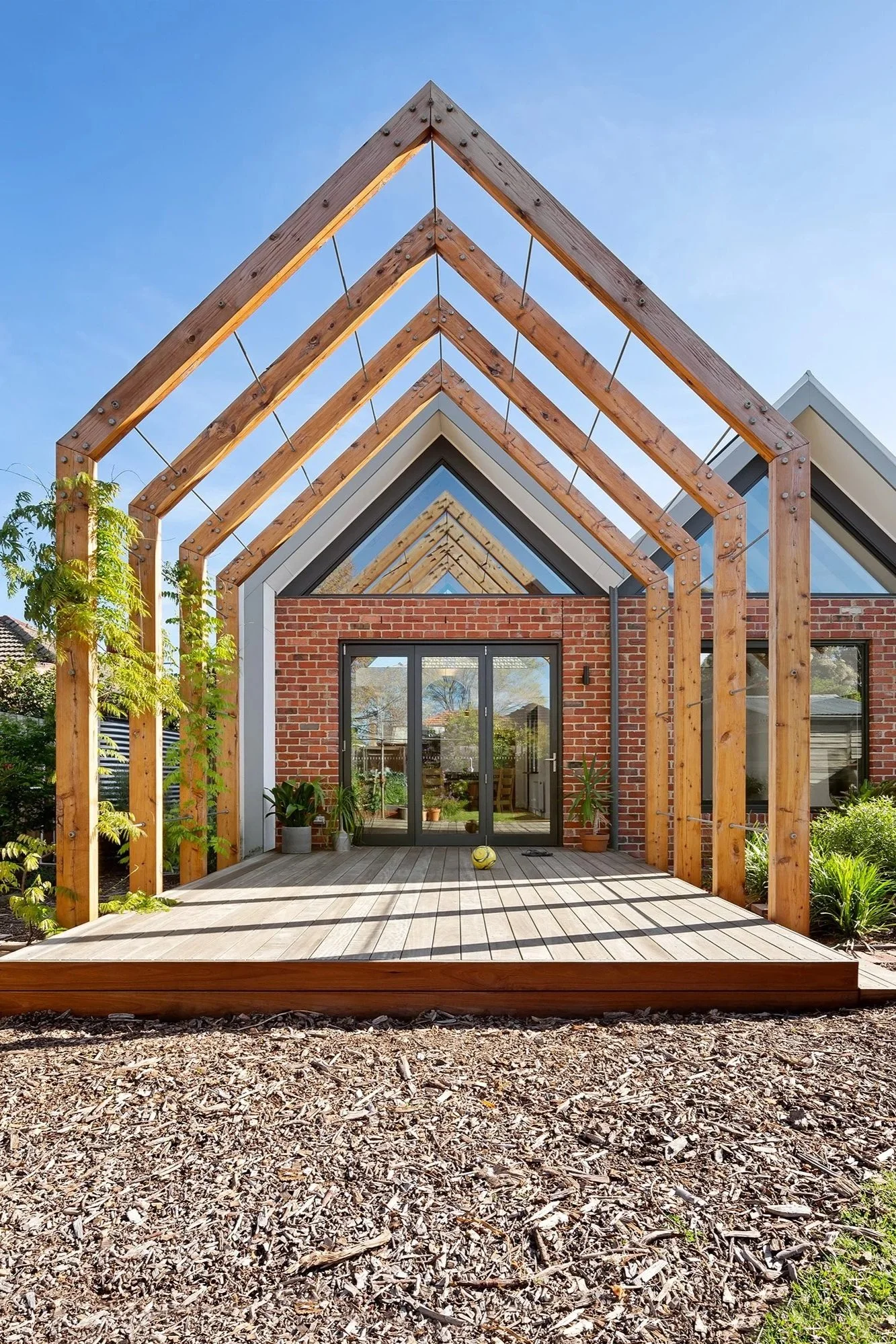 Repeated gable-form timber pergola trained with climbing vines for seasonal shading, leading to a recycled brick extension