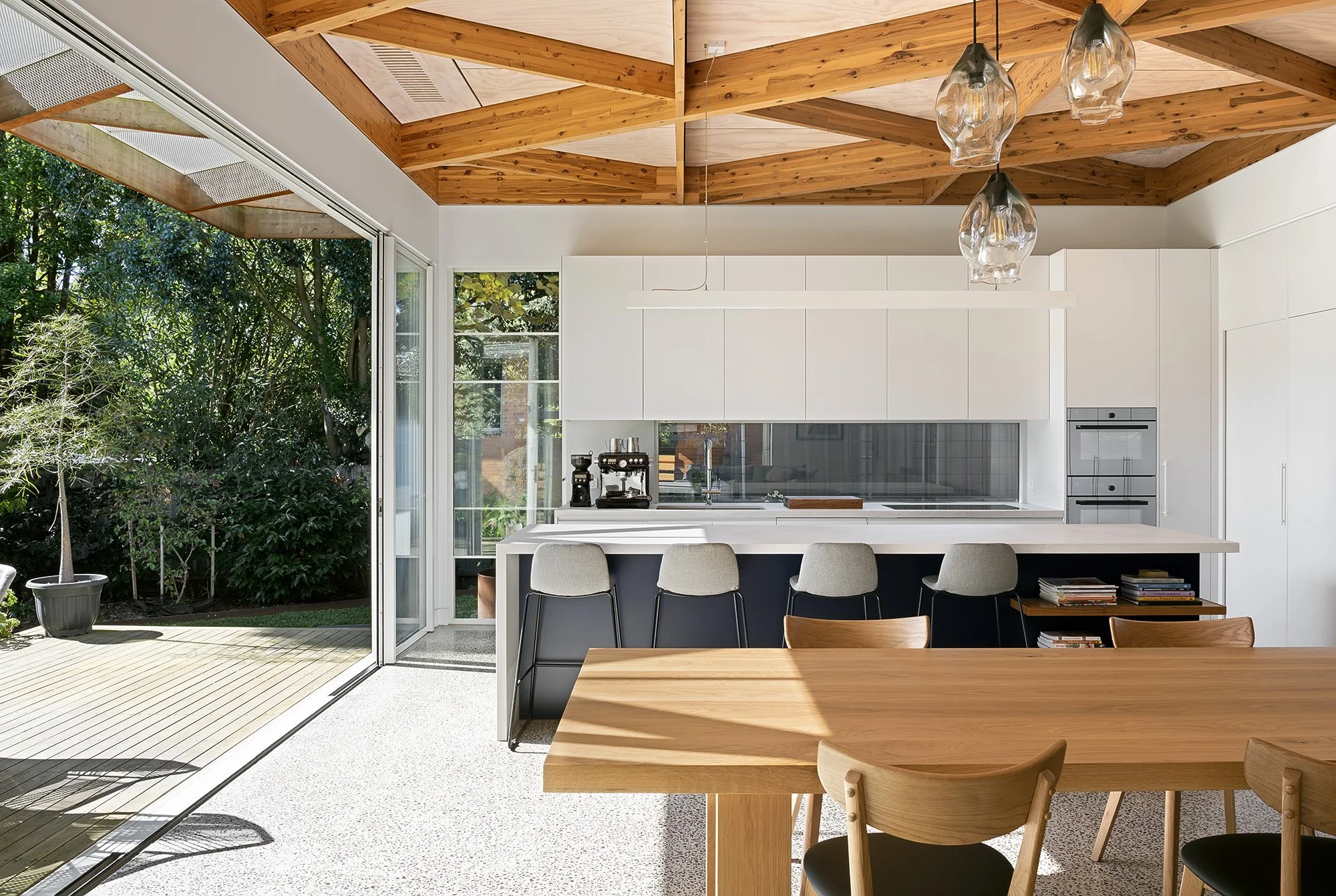 Kitchen with white cabinetry and glass pendant lighting, opening onto a garden deck beneath a framed timber ceiling