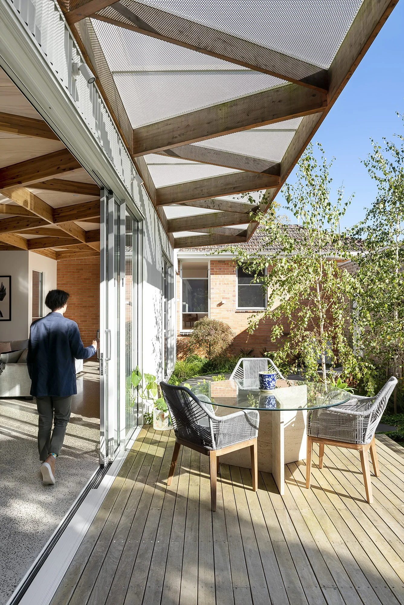 A resident opening glazed doors onto a deck shaded by a perforated steel pergola, with an outdoor dining setting beyond