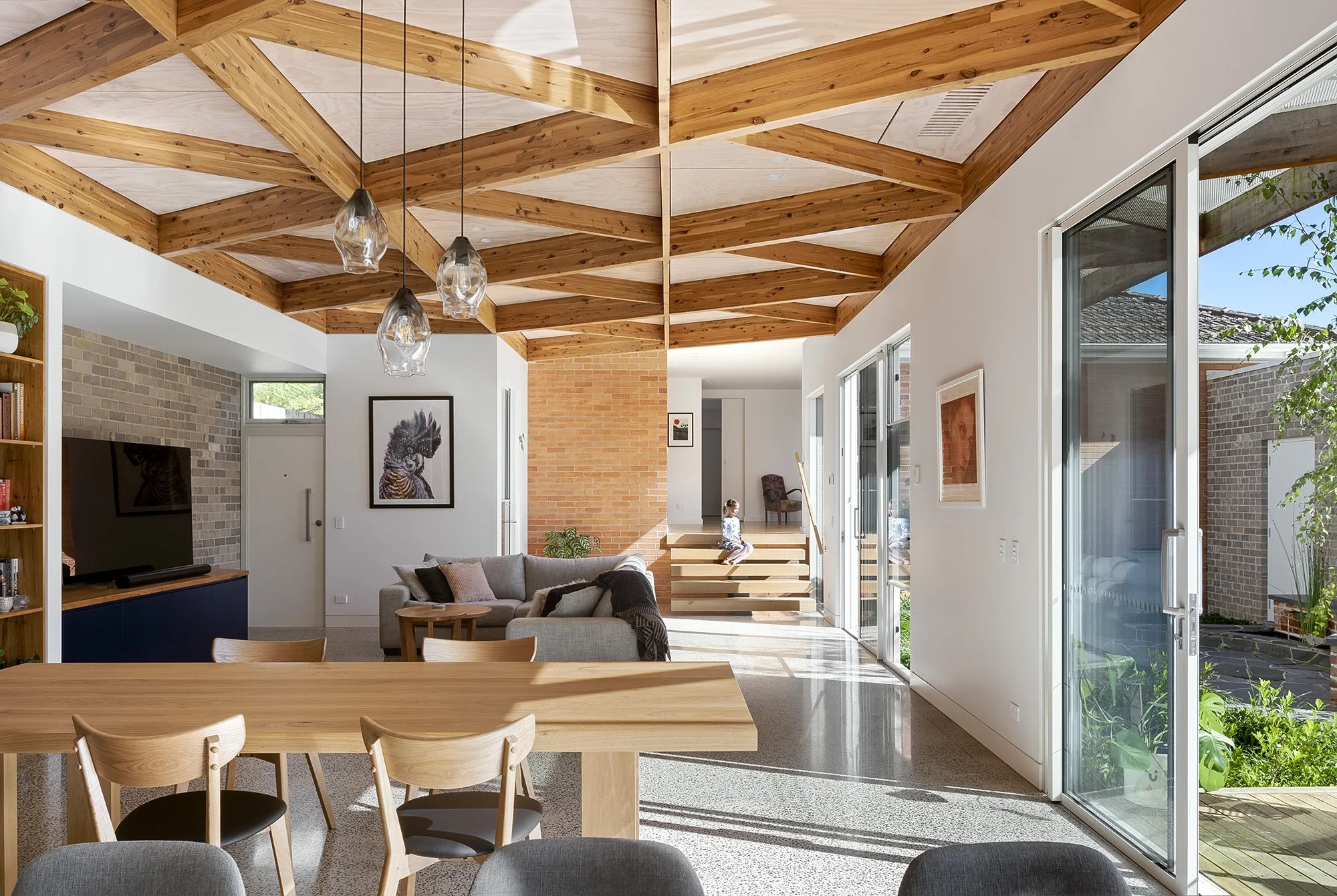 Open-plan living and dining area with a raked diagrid timber ceiling, an internal recycled brick wall and a floating timber stair
