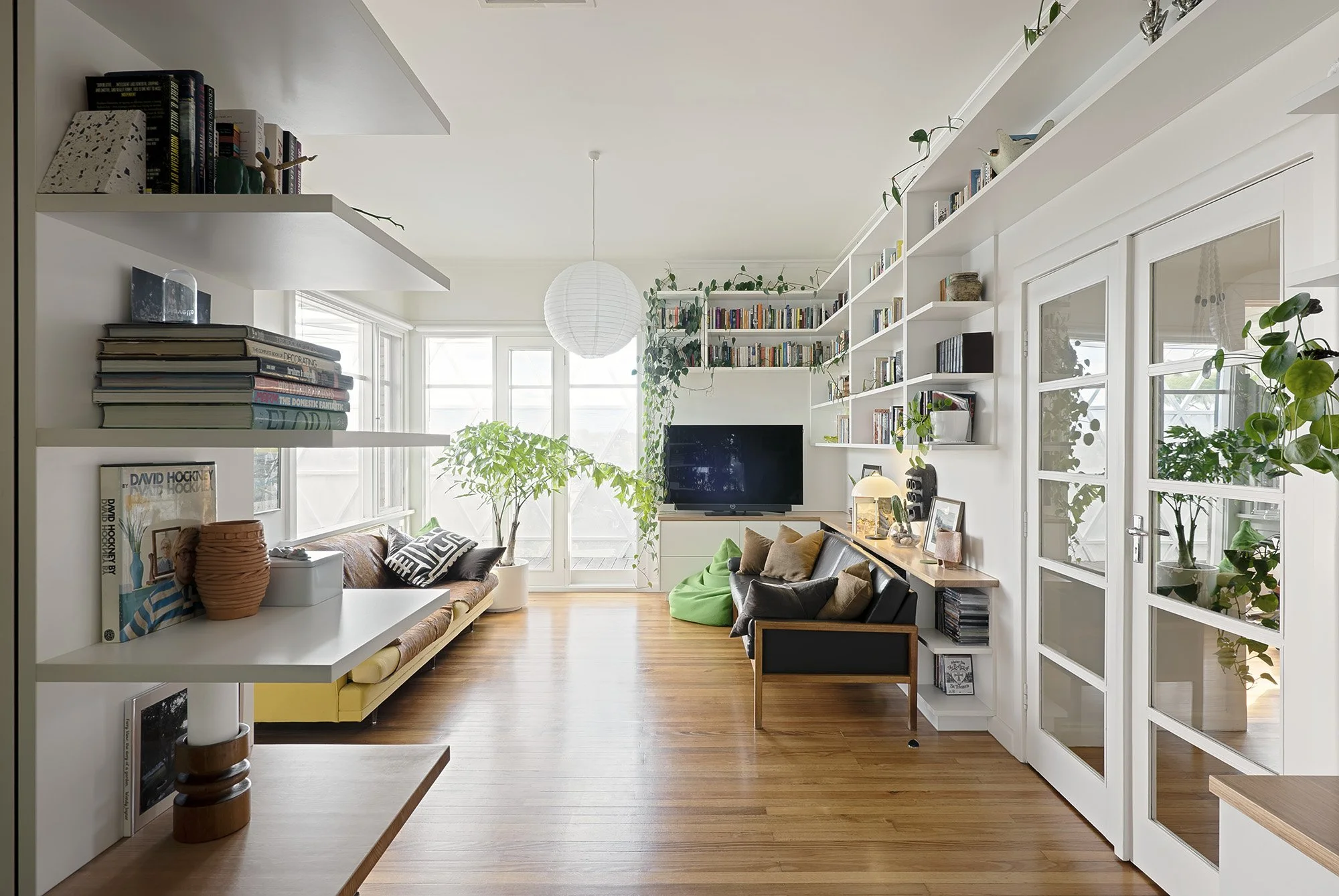 Open-plan living room with floor-to-ceiling bookshelves, timber flooring and glazed doors opening onto a leafy balcony