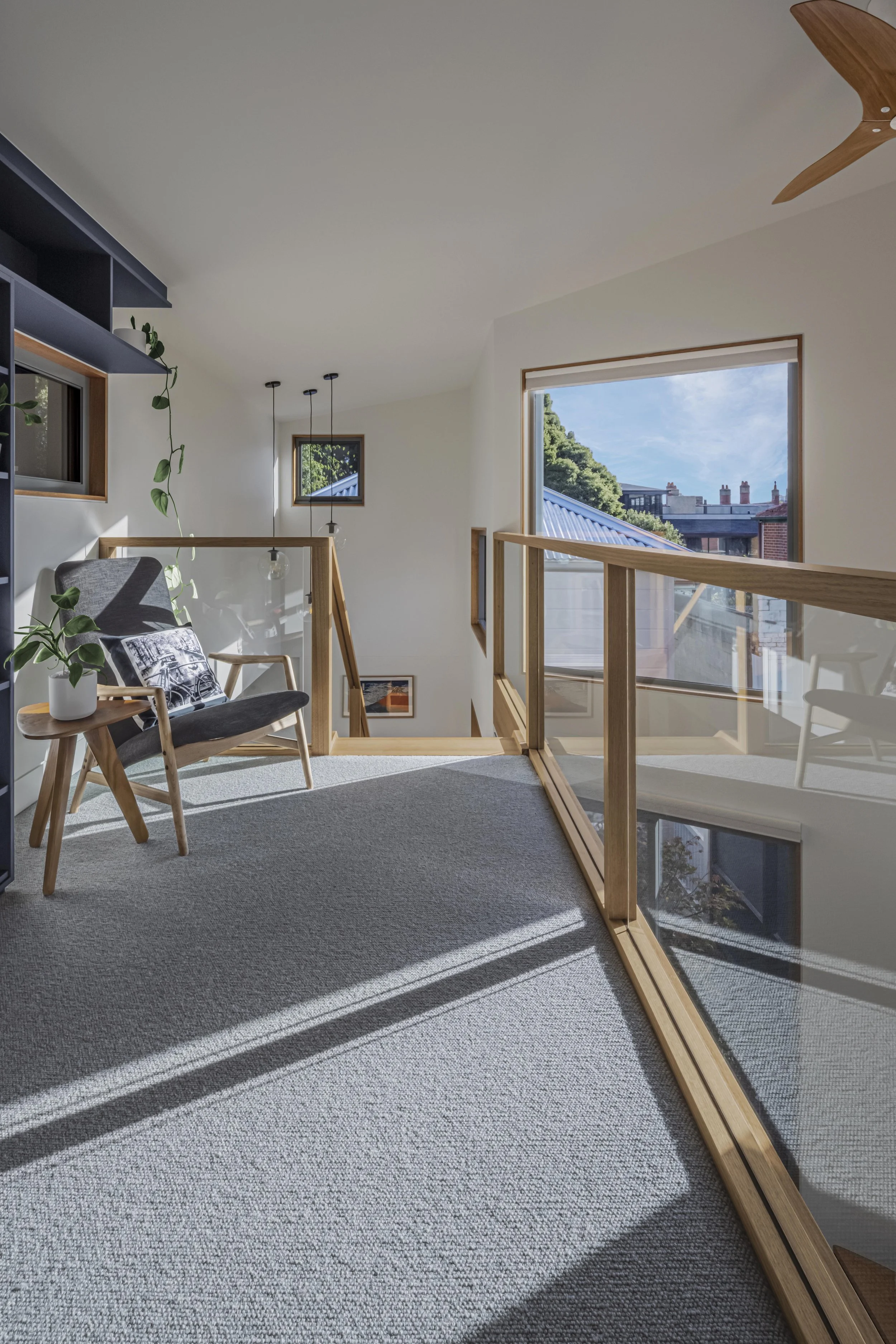 Sunlit upper-level landing with a lounge chair, glass stair balustrade and a window framing the neighbourhood's rooftops and chimneys