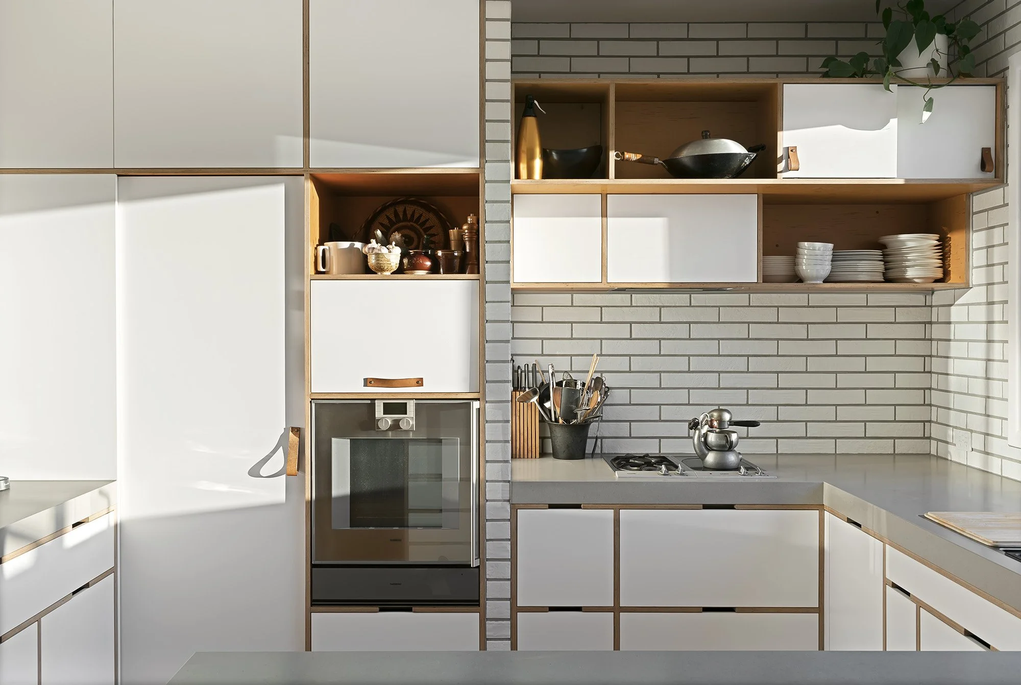 Detail of plywood kitchen joinery with open shelving, leather door pulls and a built-in oven against subway tile