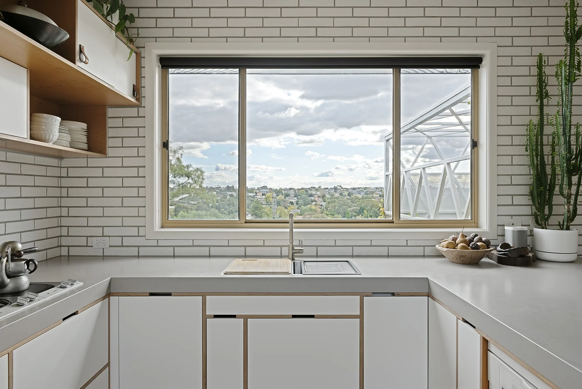 Kitchen sink positioned under a window framing treetop and rooftop views across suburban Melbourne