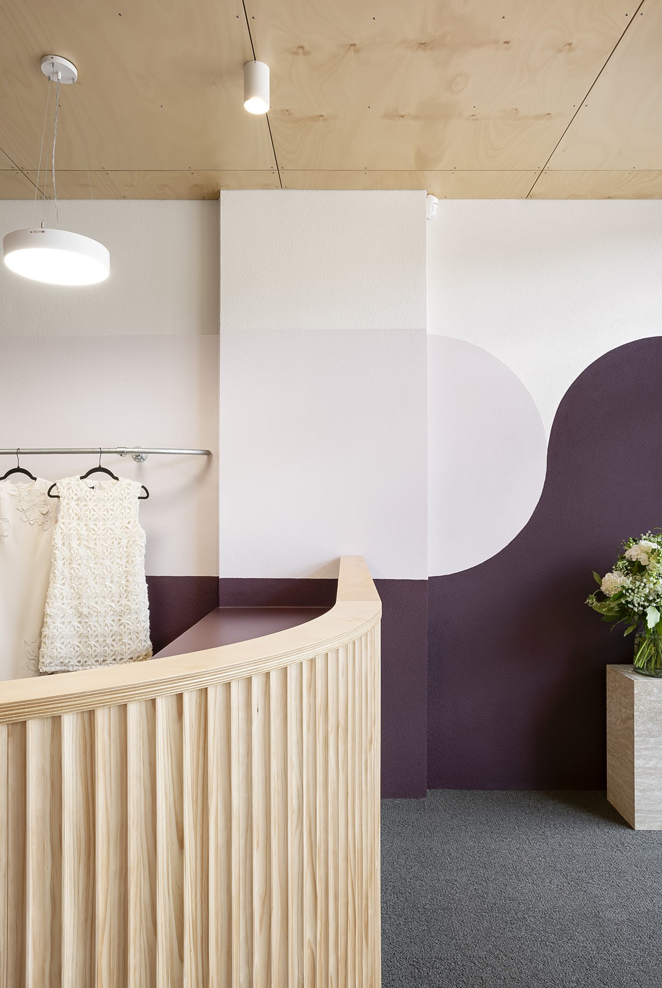 Close detail of the fluted-plywood reception desk corner, with a lace dress on display and fresh flowers on a stone plinth