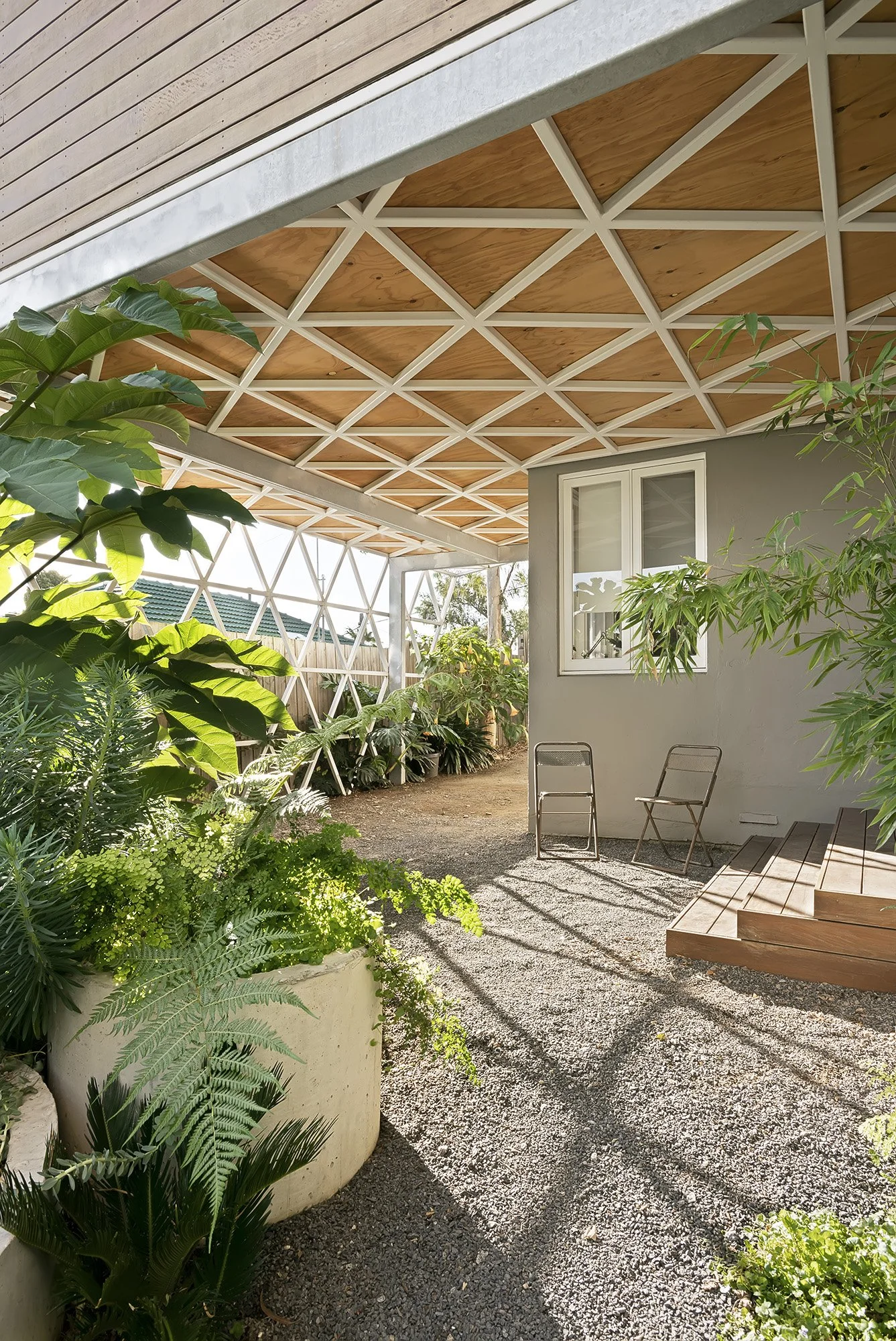 Sheltered outdoor sitting area with a geometric timber ceiling, folding chairs and dense subtropical planting