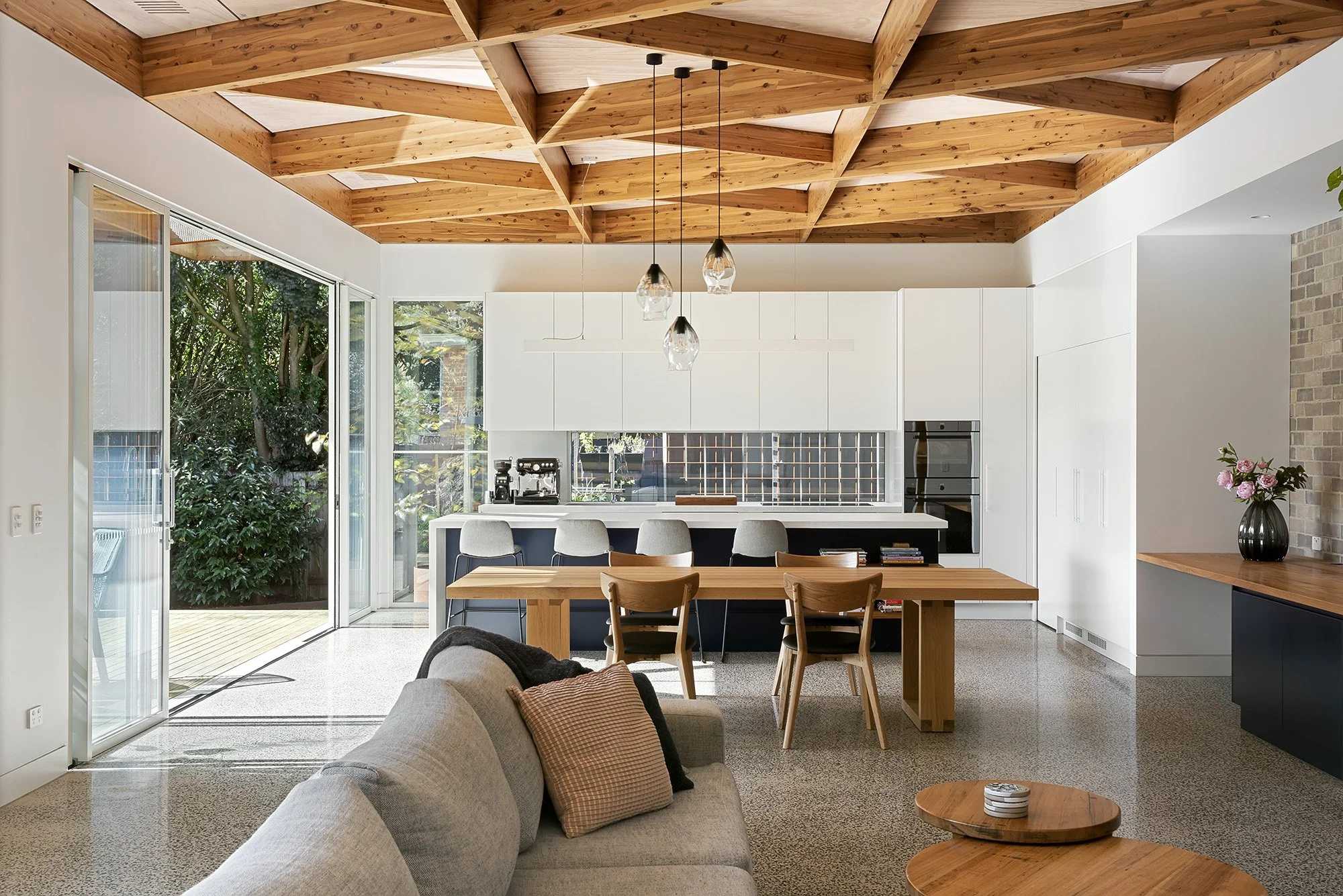Wide view of the open living, dining and kitchen space beneath the raked diagrid timber ceiling, opening onto the garden deck