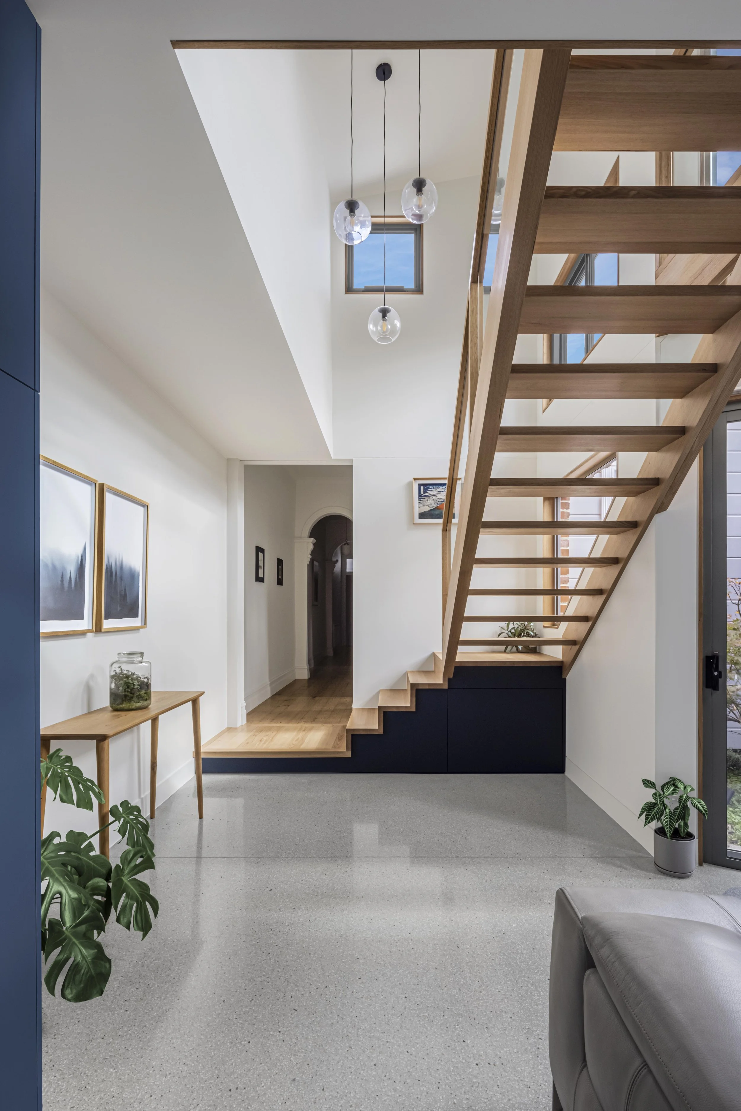Double-height stairwell with a timber and glass staircase, polished aggregate flooring and a glimpse through to the home's original arched hallway