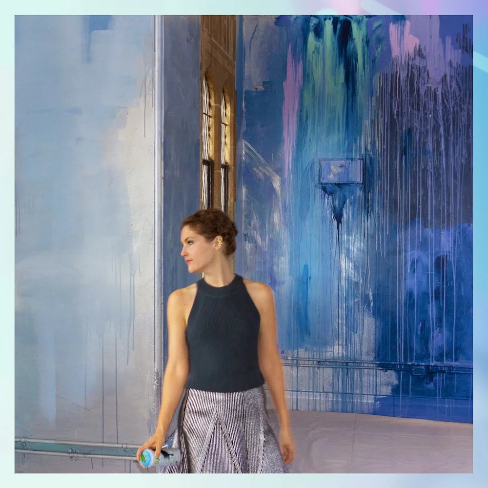 An artistic portrait of Camille Javal standing in front of one of her murals. The vibrant walls freature dripping lilac and navy paint with metallic accents..