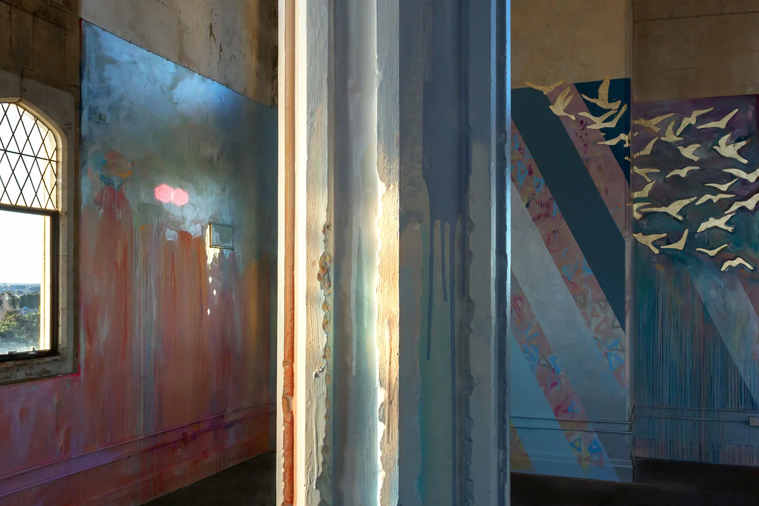 Large scale abstract mural with rainbow stripes in warehouse like tower, features gold leaf metallic birds flying out of the window, round arch windows, pillars and concrete floor.