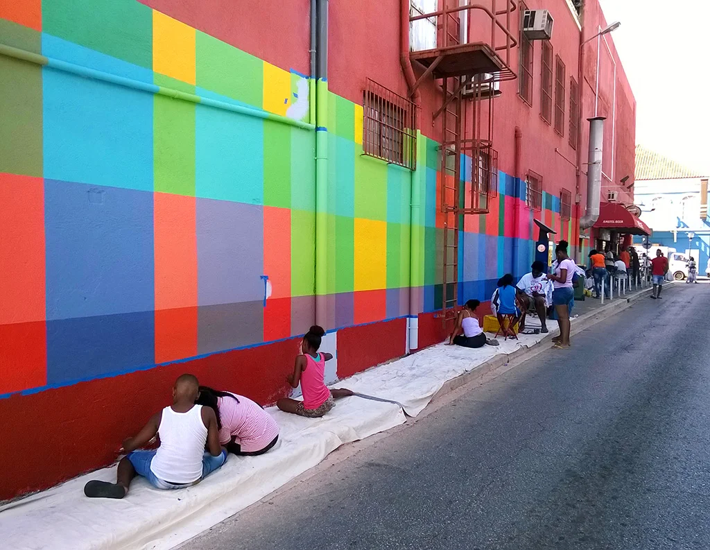 Community art mural project run by Haas & Hahn, kids help paint the design which features brightly colored geometric squares that wrap around the building like woven fabric.