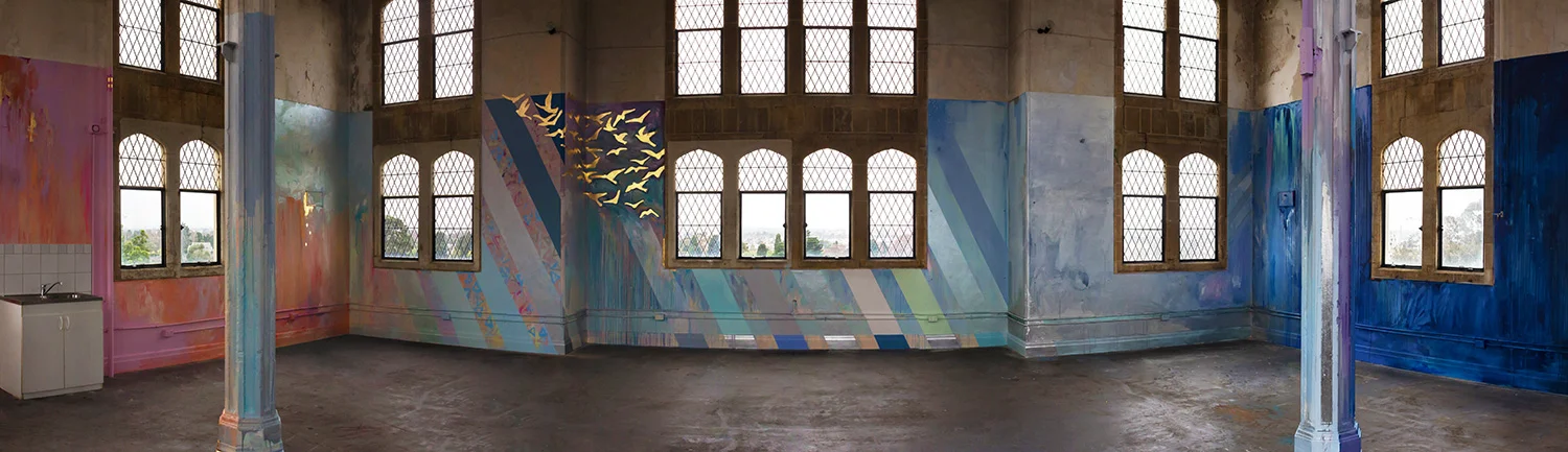 Panorama of large scale abstract mural with rainbow stripes in warehouse like tower, features gold leaf metallic birds flying out of the window, round arch windows, pillars and concrete floor.