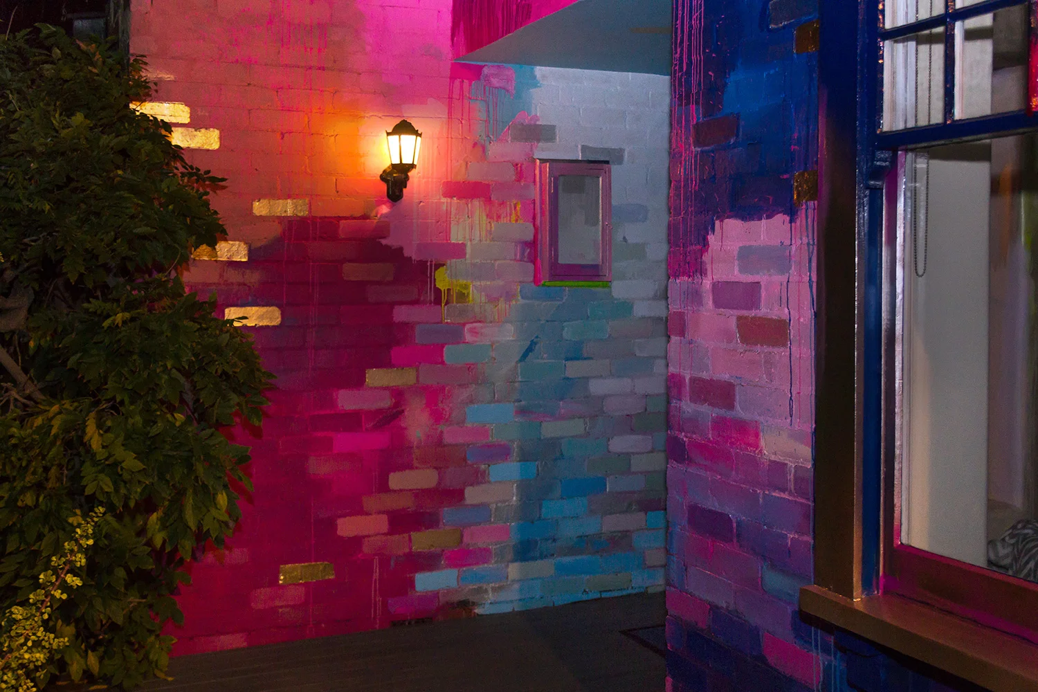 Night time glimpse at large abstract mural painted on the exterior of a house, the wall lamp lights up the metallic colored bricks.