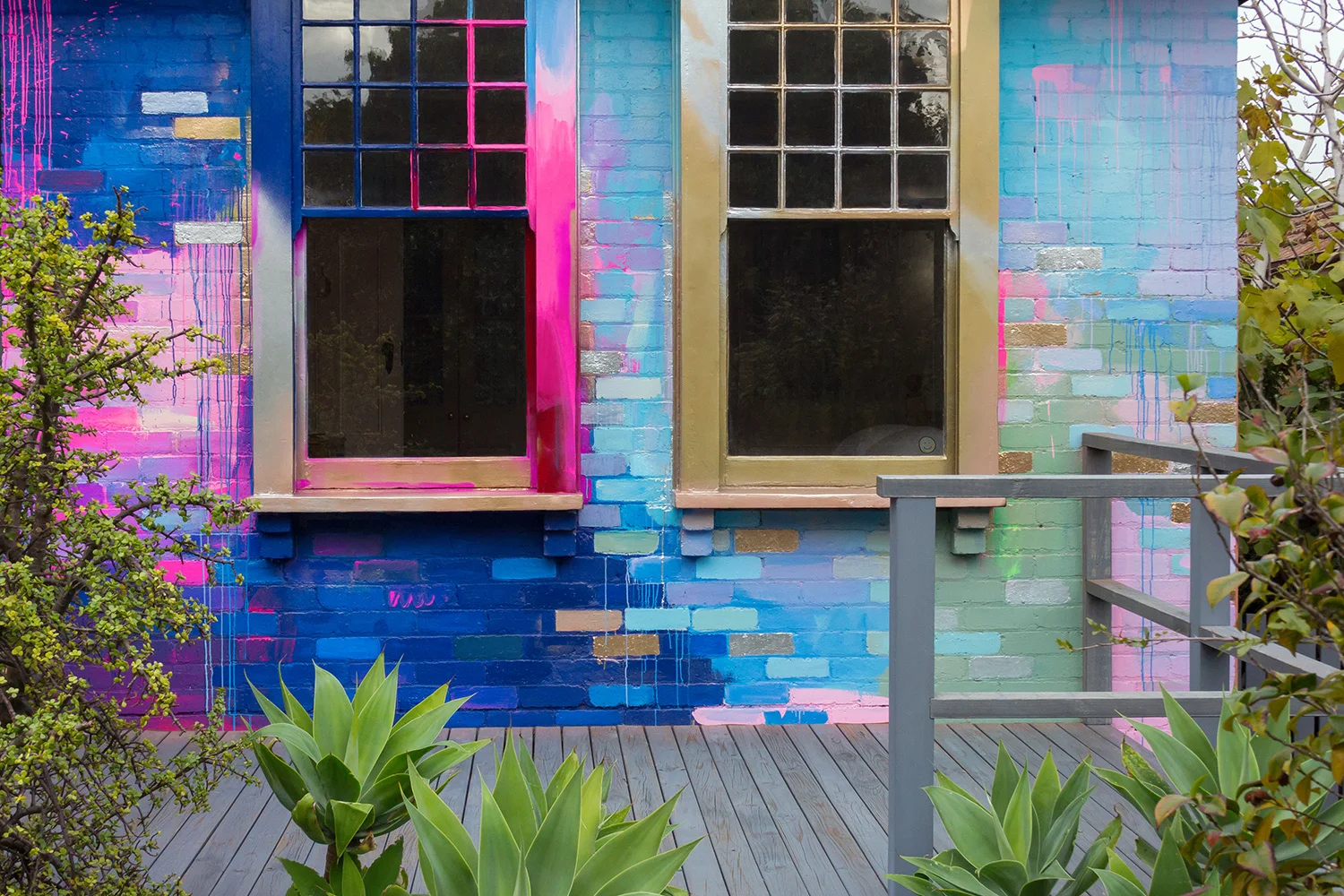 Vibrant abstract exterior mural that covers a residential home with a bright color palette of mostly pink, blue and metallic. Pops of color highlight the bricks and contrast with the garden foliage.