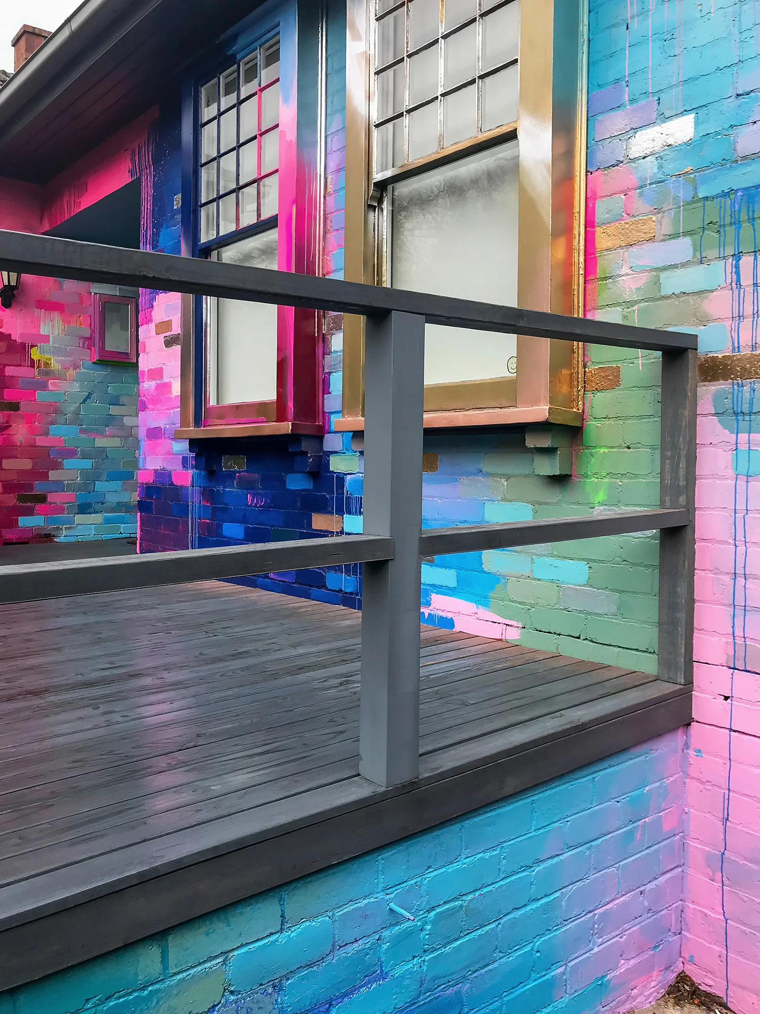 Vibrant abstract mural painted on the exterior of a house. The colors contrast with the traditional design of the house and surrounding elements. The deck, wet from rain, reflects colors from the painting.