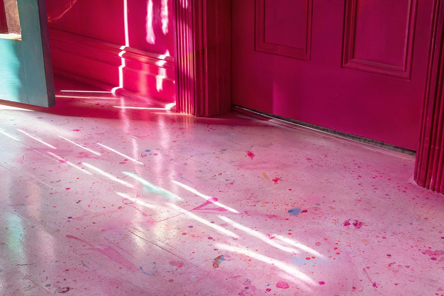Close up detail of the floor in an immersive mural. Pink paint splatters over the white wooden floor, while light pours through the door and makes striking patterns on the gloss finish.