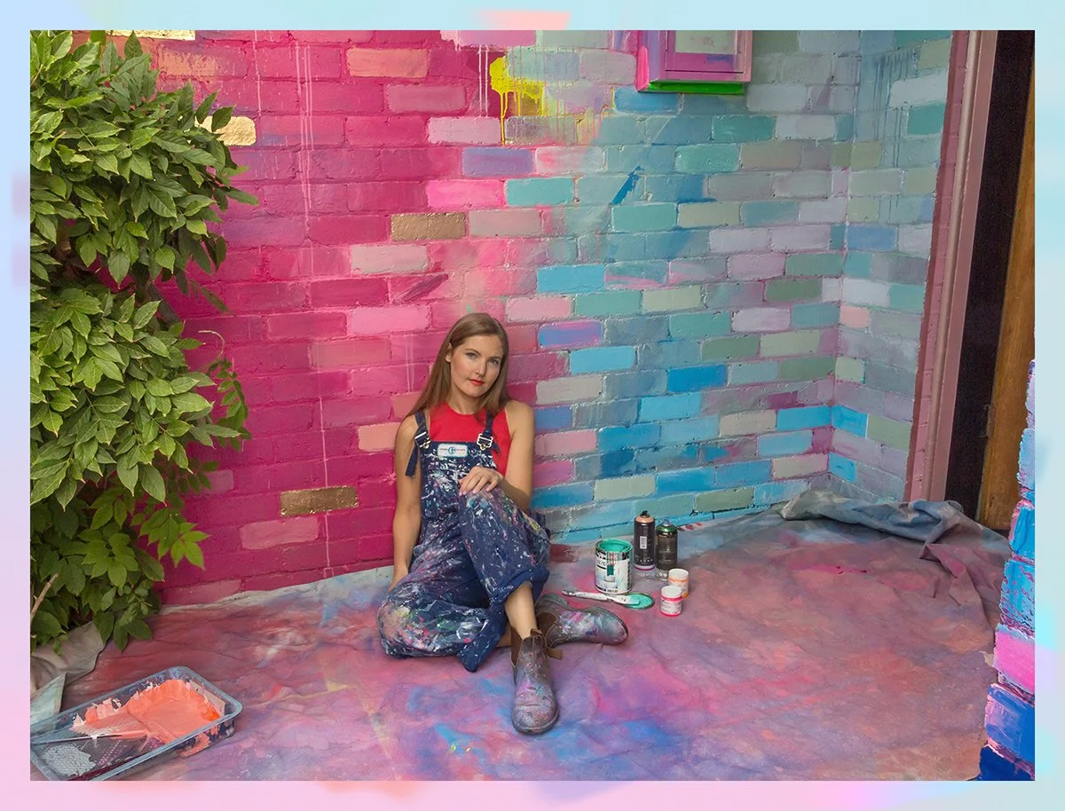 Mural artist Camille Javal leans against a brick corner wall that is dynamically painted in vibrant gradients of pink, blue, teal and purple.