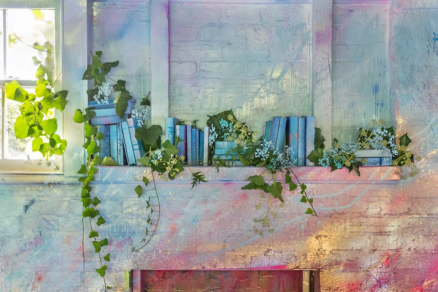 Close up of fireplace mantel, which is painted in pink, blue and gold brushstrokes. Blue books line the mantel while foliage grows between them.