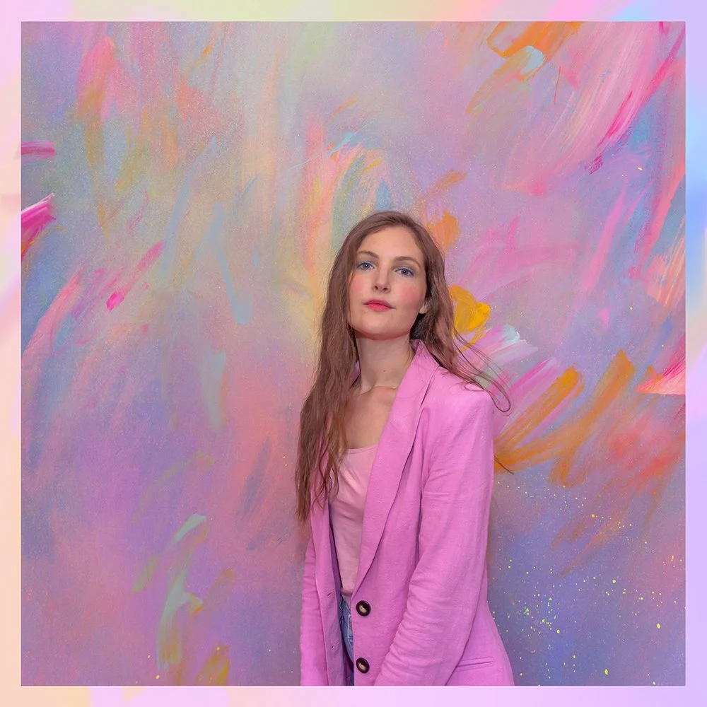 Artist Camille Javal wearing a pink blazer stands in front of a colorful, abstract pastel mural with brushstrokes of pink, purple and yellow.
