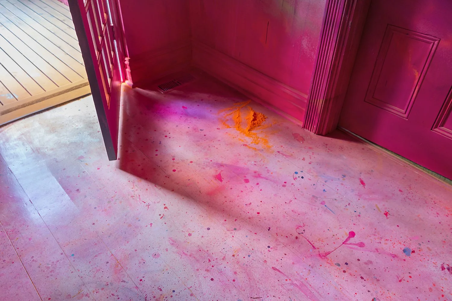 The painted floor of an immersive mural, featuring pink splatters over a white wooden floor. Yellow powder from a smoke cannon has been swept into a pile.