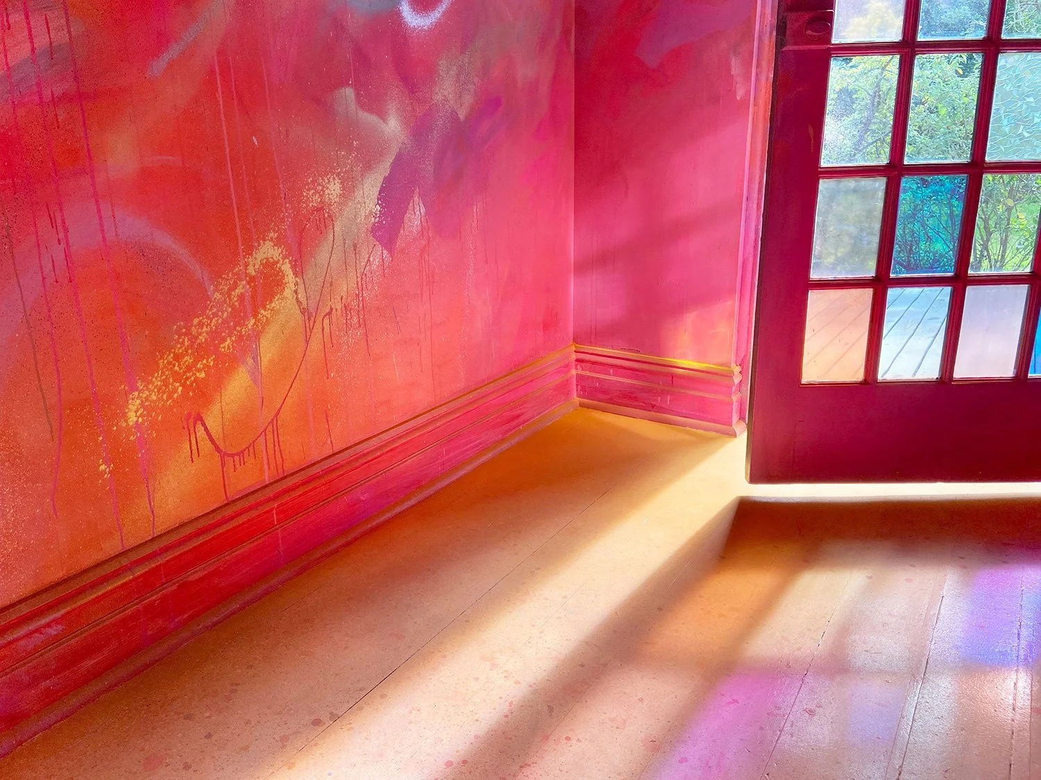 Light shines through a glass front door, highlighting yellow powder from a smoke bomb on the ground. The walls of the art installation are painted red with gold and pink brushmarks.