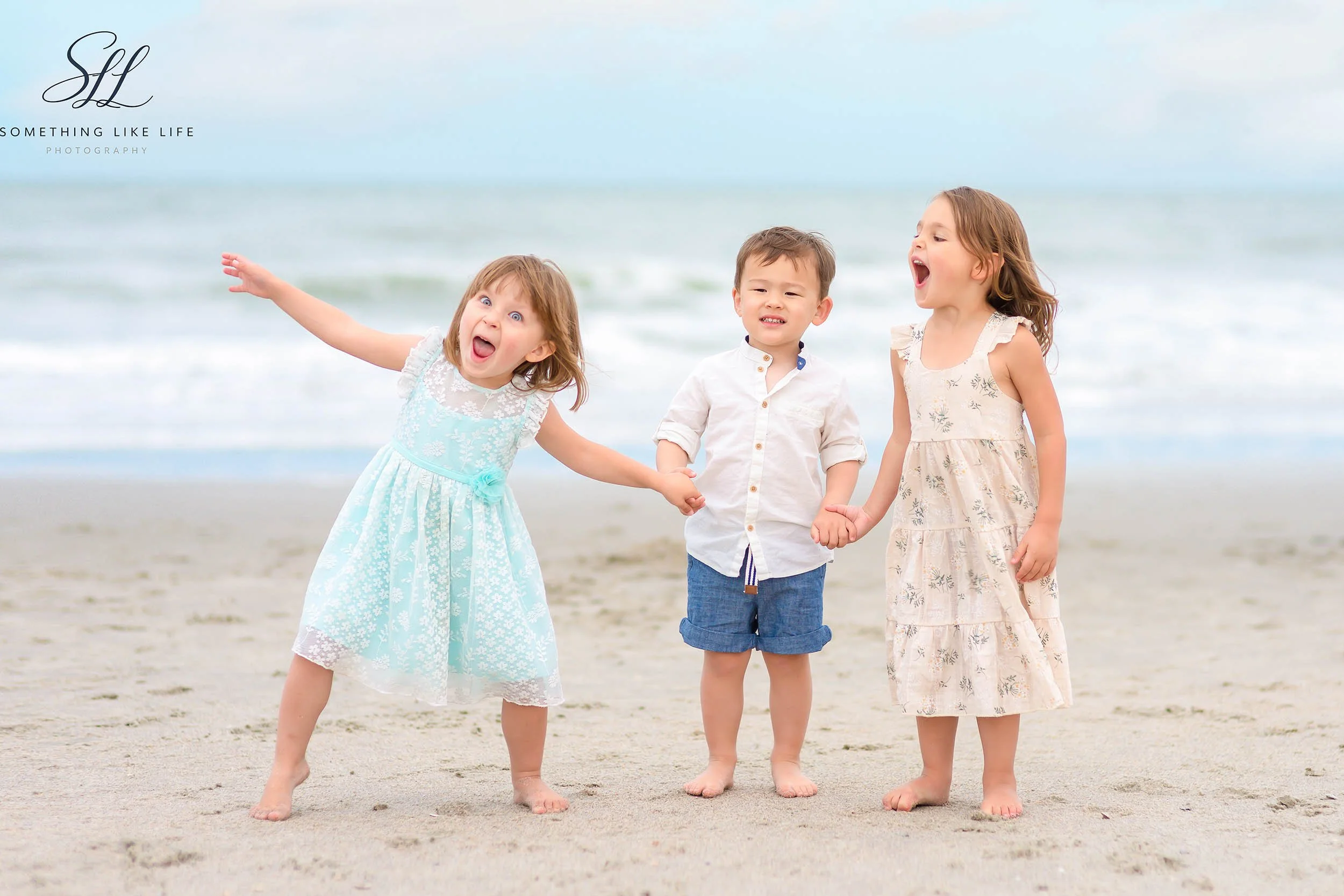 Three young children laughing and holding hands during a candid beach session on their Myrtle Beach vacation, captured with soft ocean tones in the background.