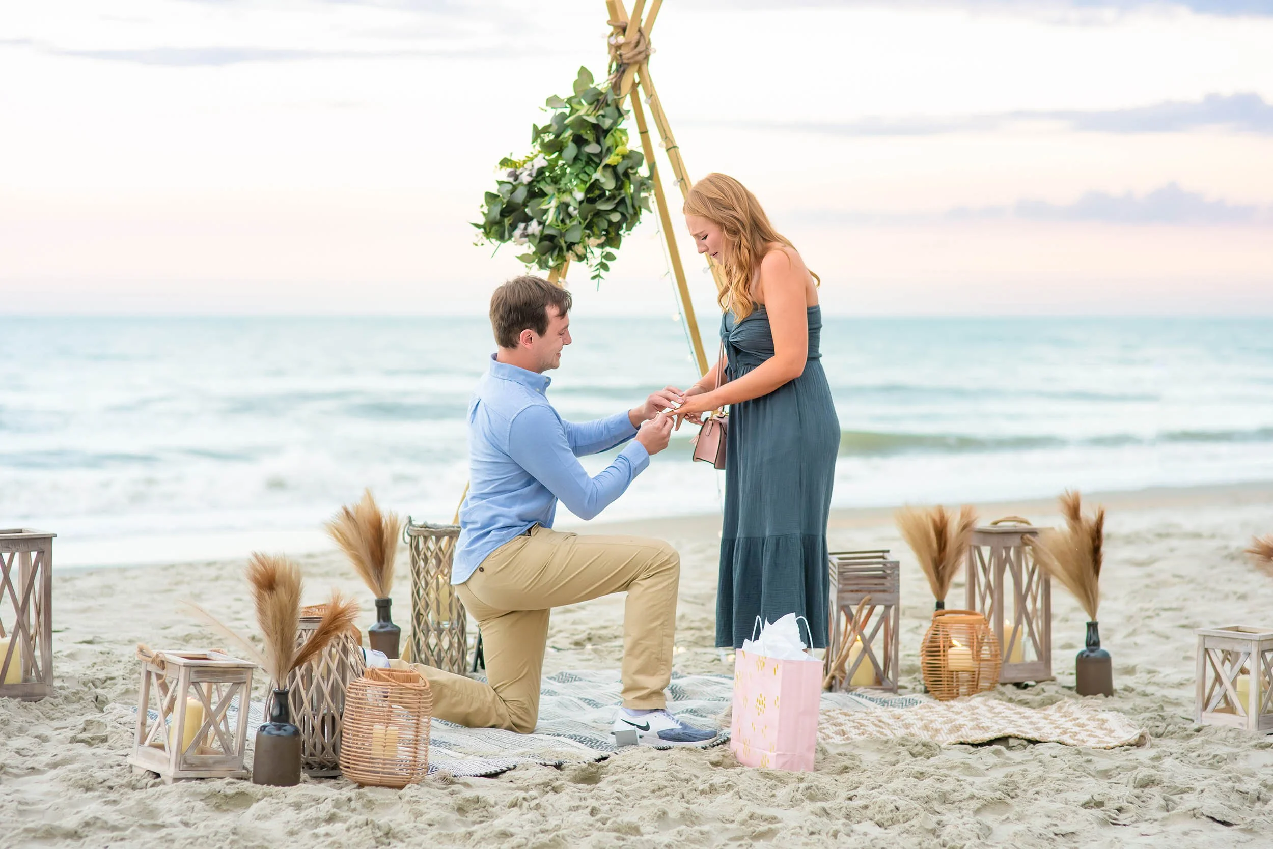 A man kneels to propose to his partner on the beach in Pawleys Island, South Carolina, with a decorated arch and ocean backdrop during a surprise engagement session.