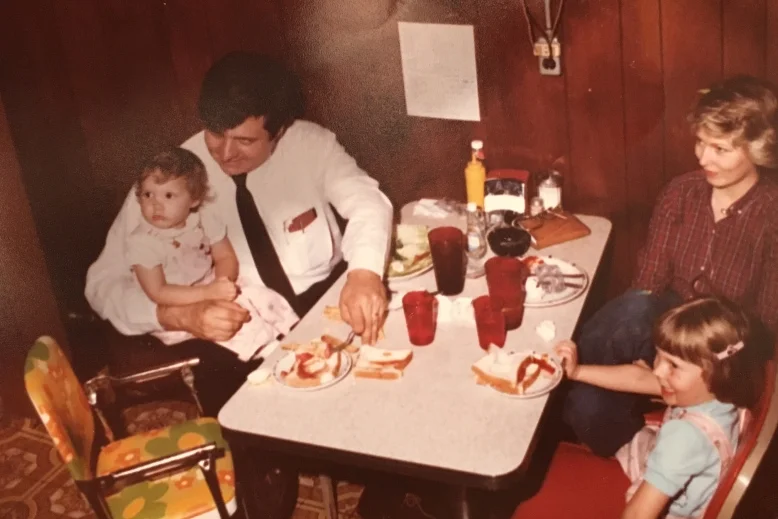 Me and my sister in matching overalls eating with my parents. Check out that floral covered high chair!