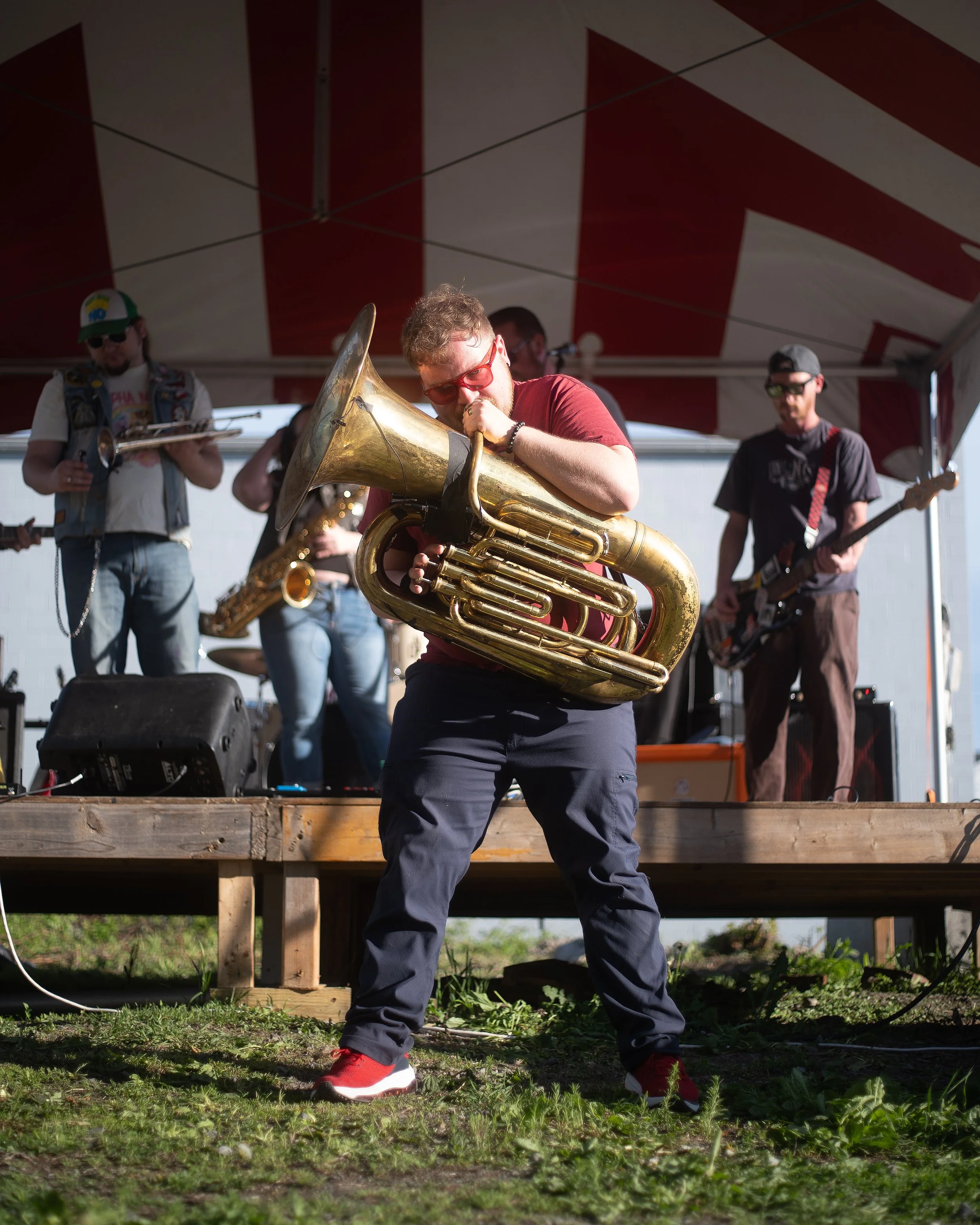 A band performing under an outdoor tent, with a musician playing a sousaphone in the foreground, wearing glasses and a red shirt, with other band members in the background including a guitarist and a trombone player.