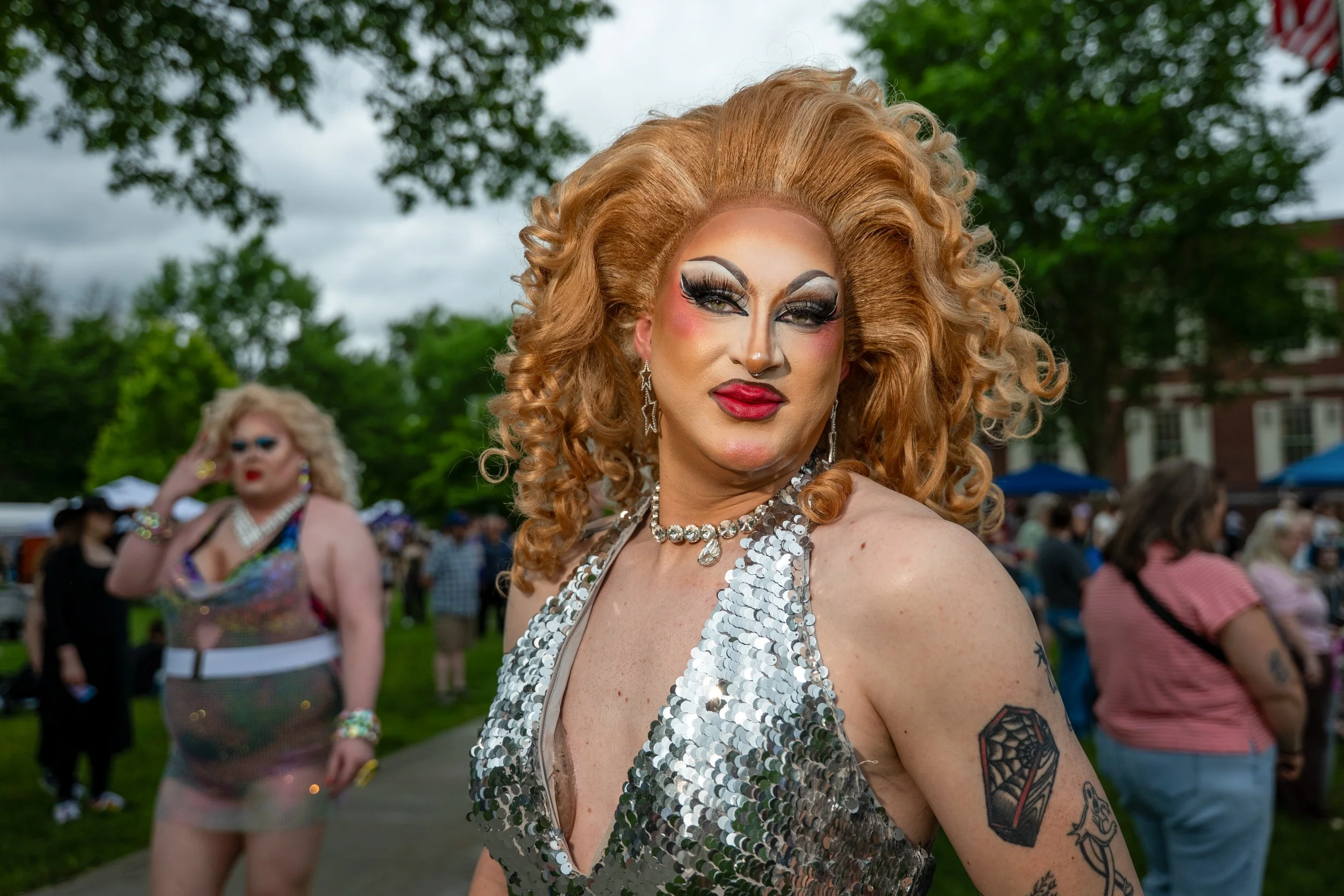 A person with curly blonde hair styled in voluminous waves, wearing dramatic makeup with bold eye makeup, pink blush, and red lipstick, dressed in a silver sequin top, with tattoos on their arm, at an outdoor event with trees, people, and tents in th