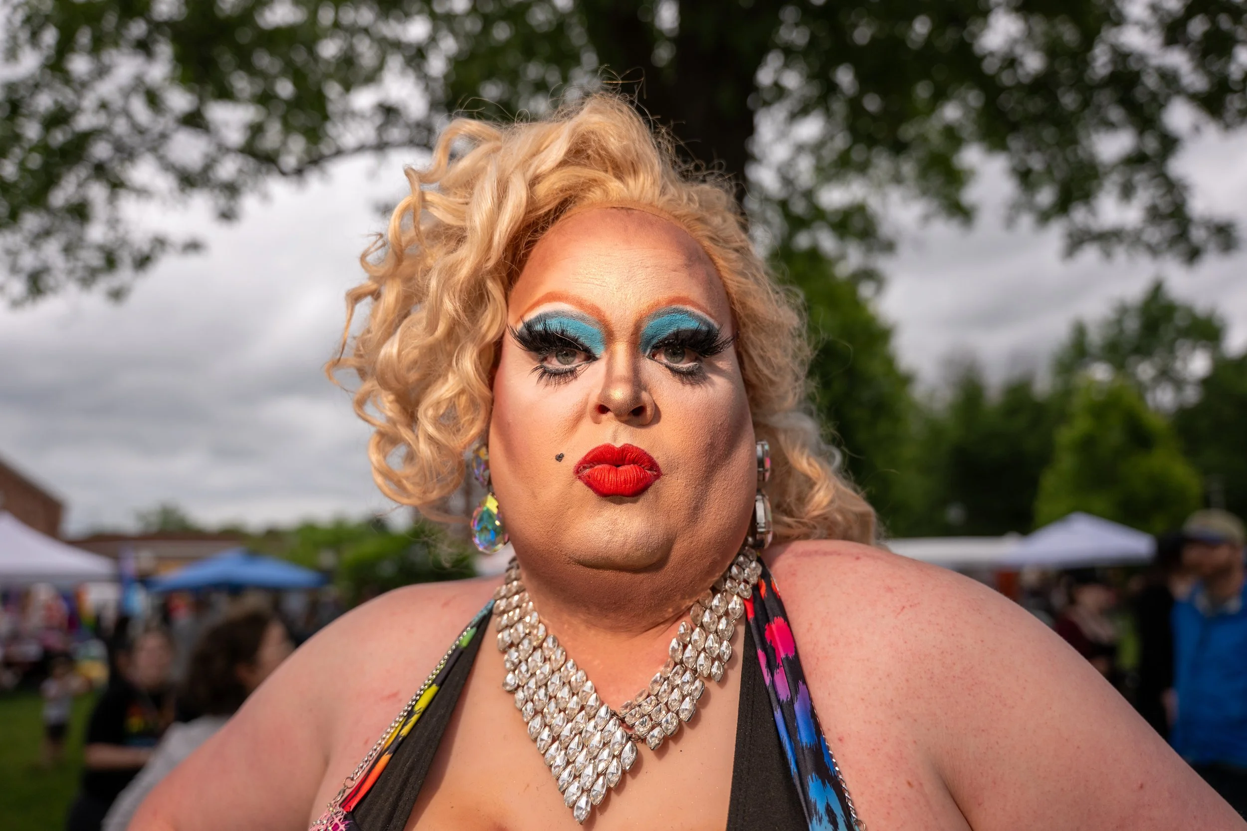 Close-up of a person with blonde curly hair and dramatic makeup, wearing large earrings, a colorful top, and layered necklaces, outdoors at a crowded event under cloudy skies.