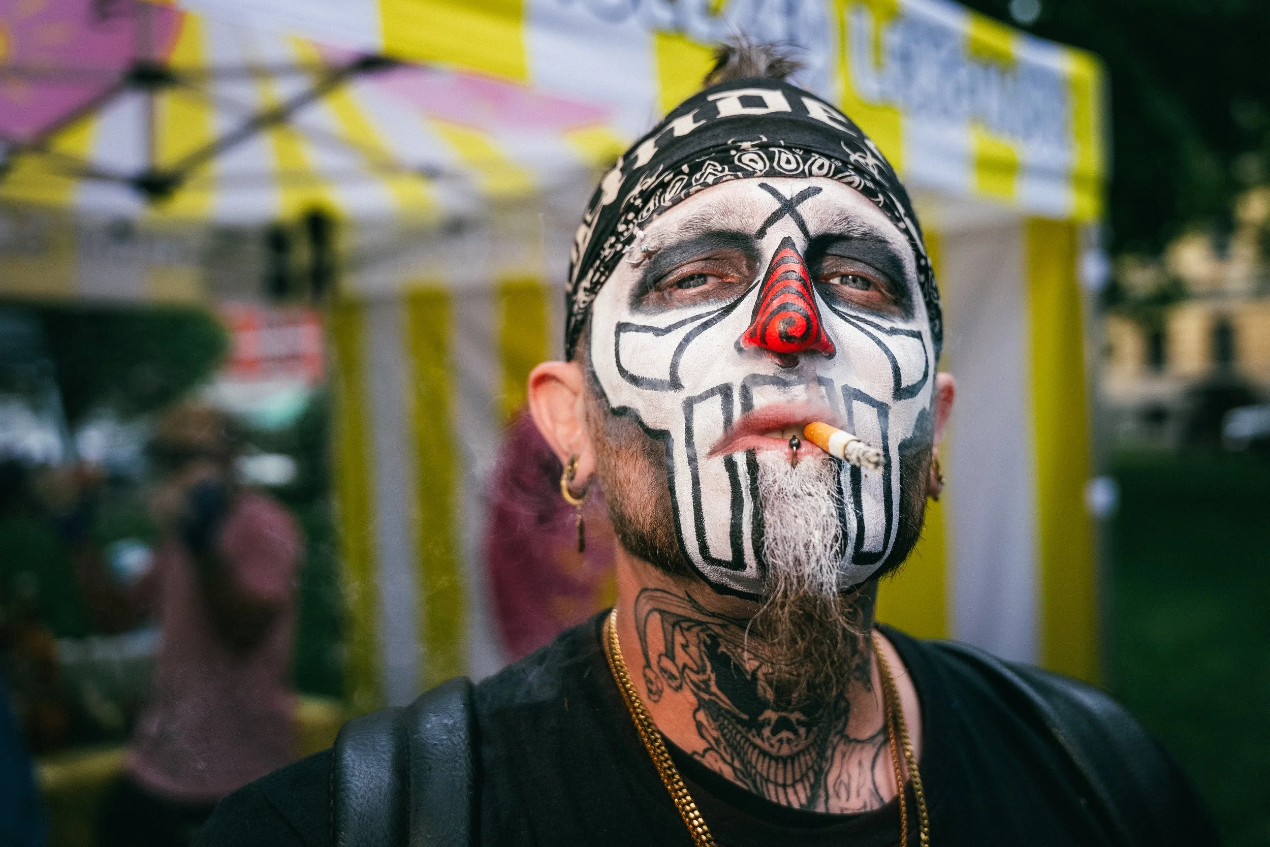 Close-up of a man wearing face paint resembling a skull, with a red nose and black markings, smoking a cigarette, wearing a black leather jacket, gold chain, and black bandana with white patterns, in an outdoor setting.