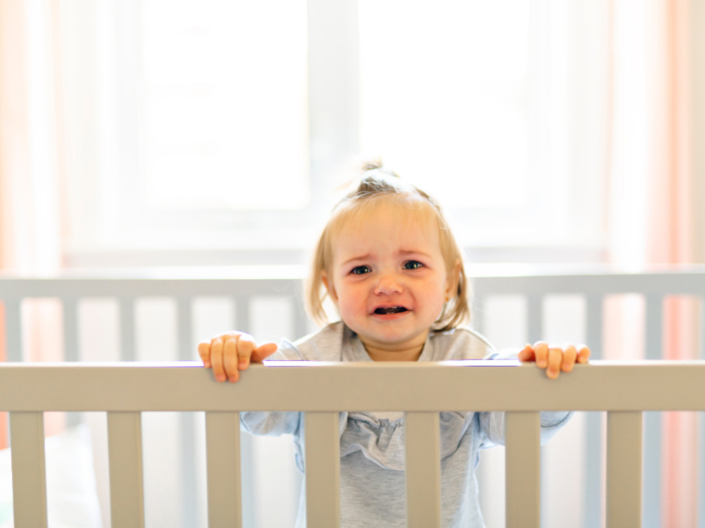 Toddler girl with light hair stands in her crib, holding onto the railing and frowning at the camera, her face verging on tears.