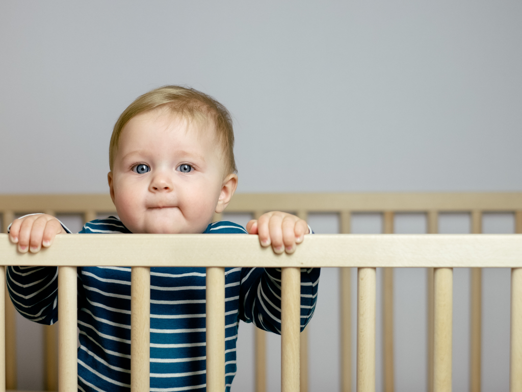 Baby boy uses the crib railing to pull up, looking out towards the camera. His eyes are wide open as if he's avoiding falling asleep.