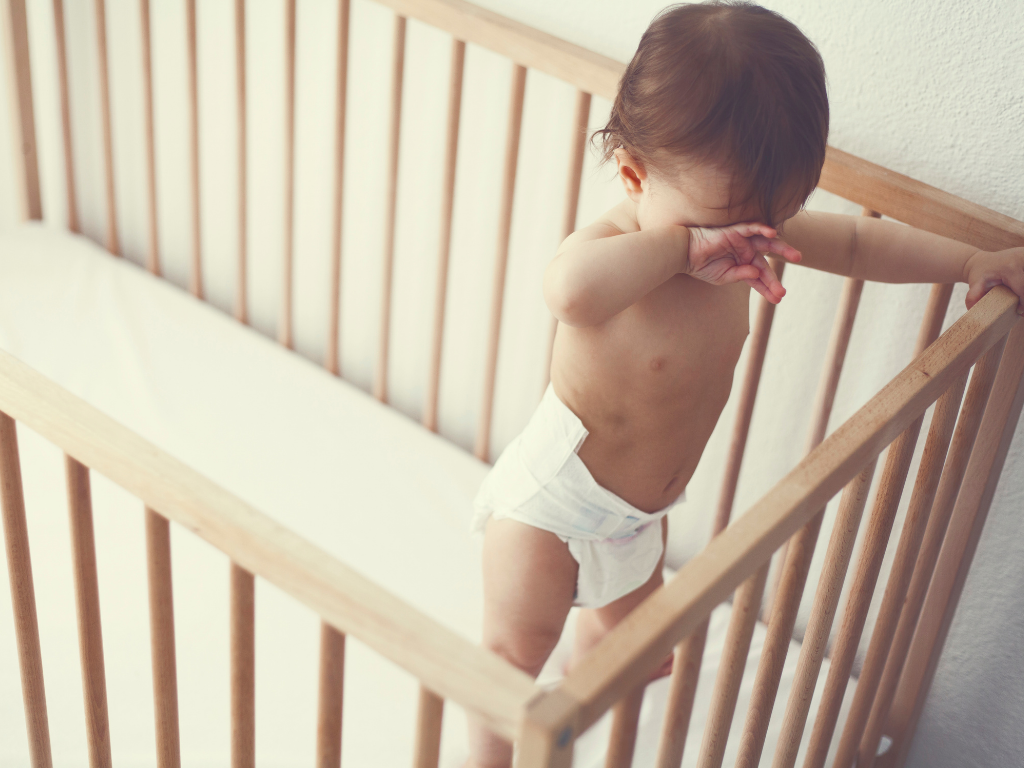 Older baby stands up in their crib, looking down towards their toes and rubbing their eyes as if sleepy or upset.