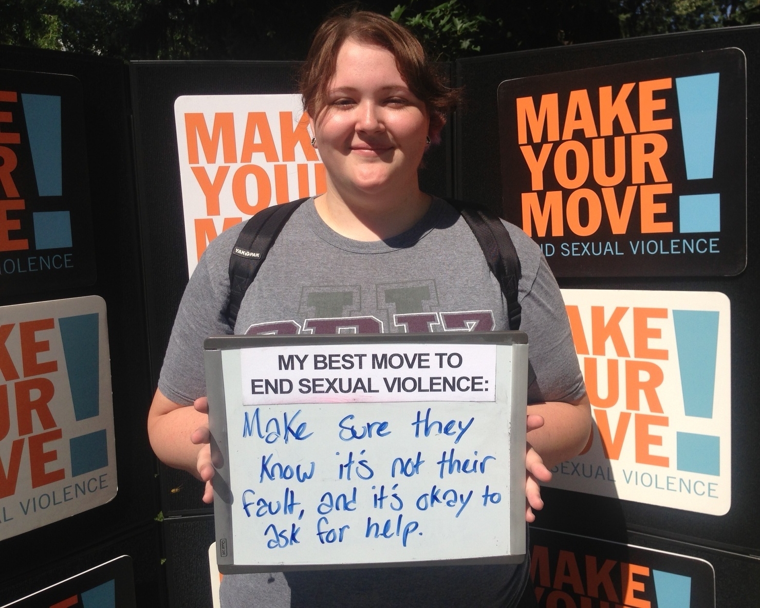 A person with brown hair holds a whiteboard. The sign says: "My best move to end sexual violence: Make sure they know it's not their fault, and it's okay to ask for help.”