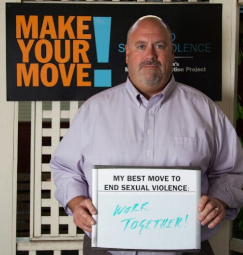 A man stands in from of a Make Your Move! sign. He is holding a whiteboard that reads “My best move to end sexual violence: Work Together!”
