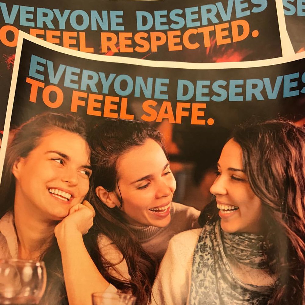 A pile of patron posters show three women at a bar laughing together. The header reads “Everyone deserves to feel safe.”