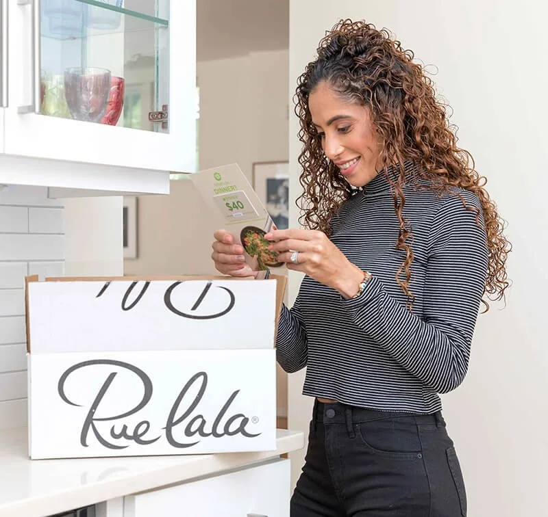 Person in a kitchen holding a package insert card while unpacking a white Rue La La box on the counter.
