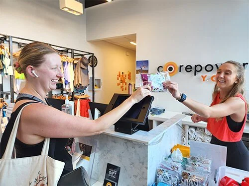 Lifestyle brand sampling moment with a packaged item being handed across a checkout counter, surrounded by merchandise displays.