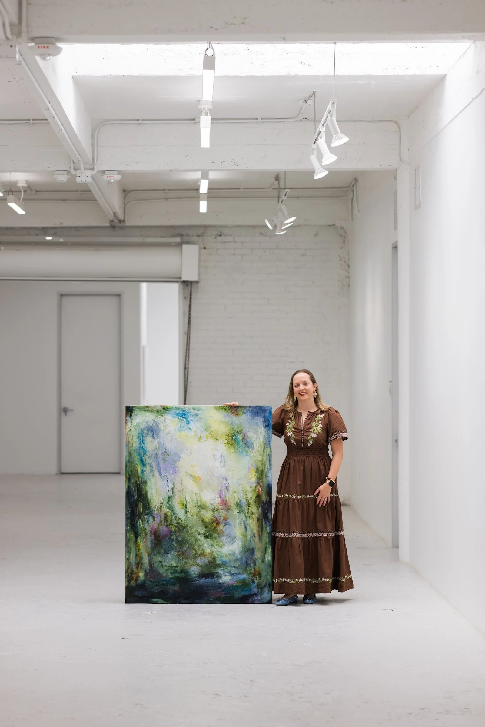 Abstract artist Allyson Caire pictured in the Winter Street Studios gallery space, smiling, next to a large abstract painting with green, blue, yellow, and purple colors.