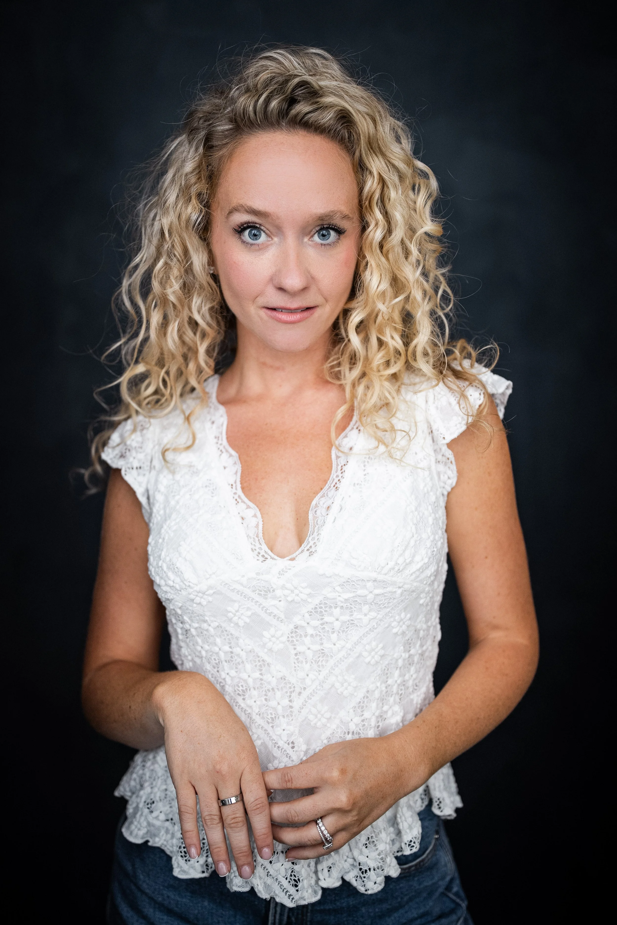 Maggie Bera. A woman with blonde, curly hair and blue eyes wearing a white lace top and blue jeans, holding her hands together in front of her against a dark background.