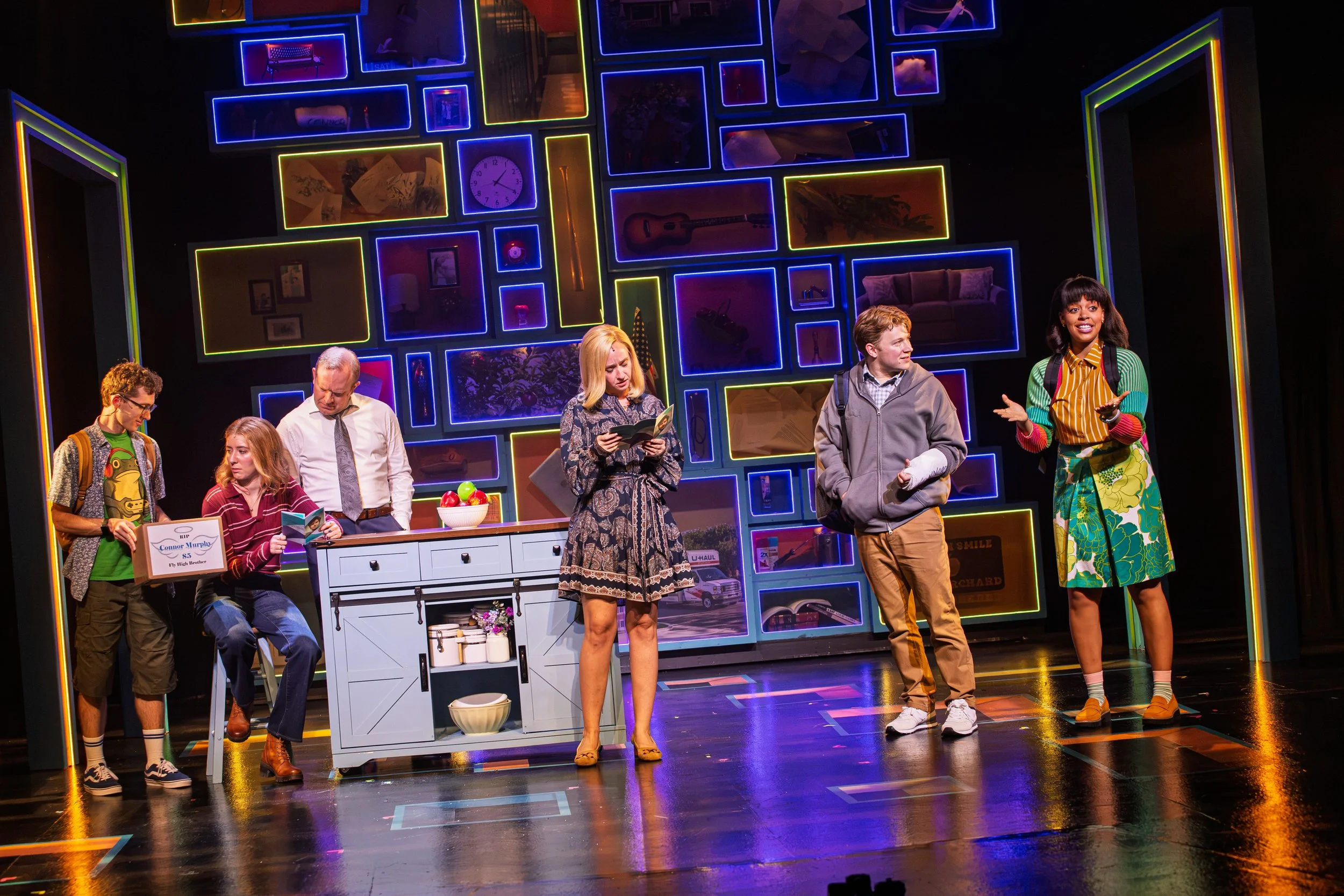 The image depicts a scene from a theatrical play with five actors on stage. The stage background is decorated with illuminated picture frames containing various objects and images.Maggie Bera playing Cynthia Murphy in Dear Evan Hansen at the Engeman 