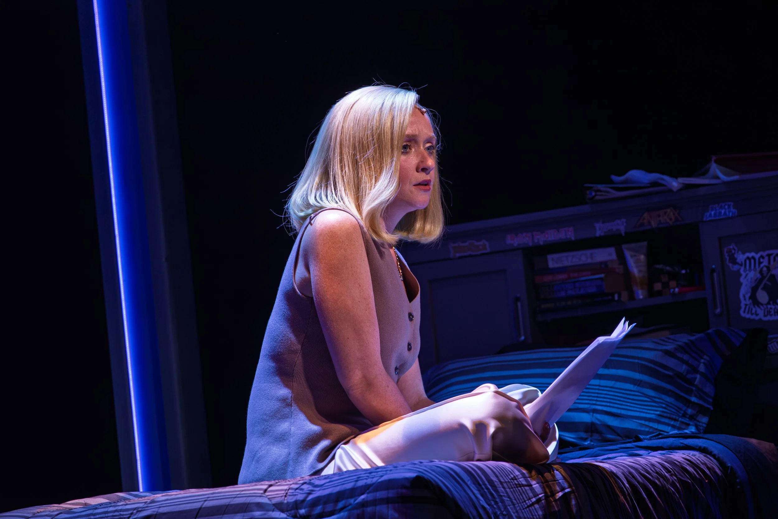 A woman with blonde hair sitting on a bed, reading from a piece of paper, in a dimly lit room with neon blue lighting. Maggie Bera playing Cynthia Murphy in Dear Evan Hansen at the Engeman Theatre.