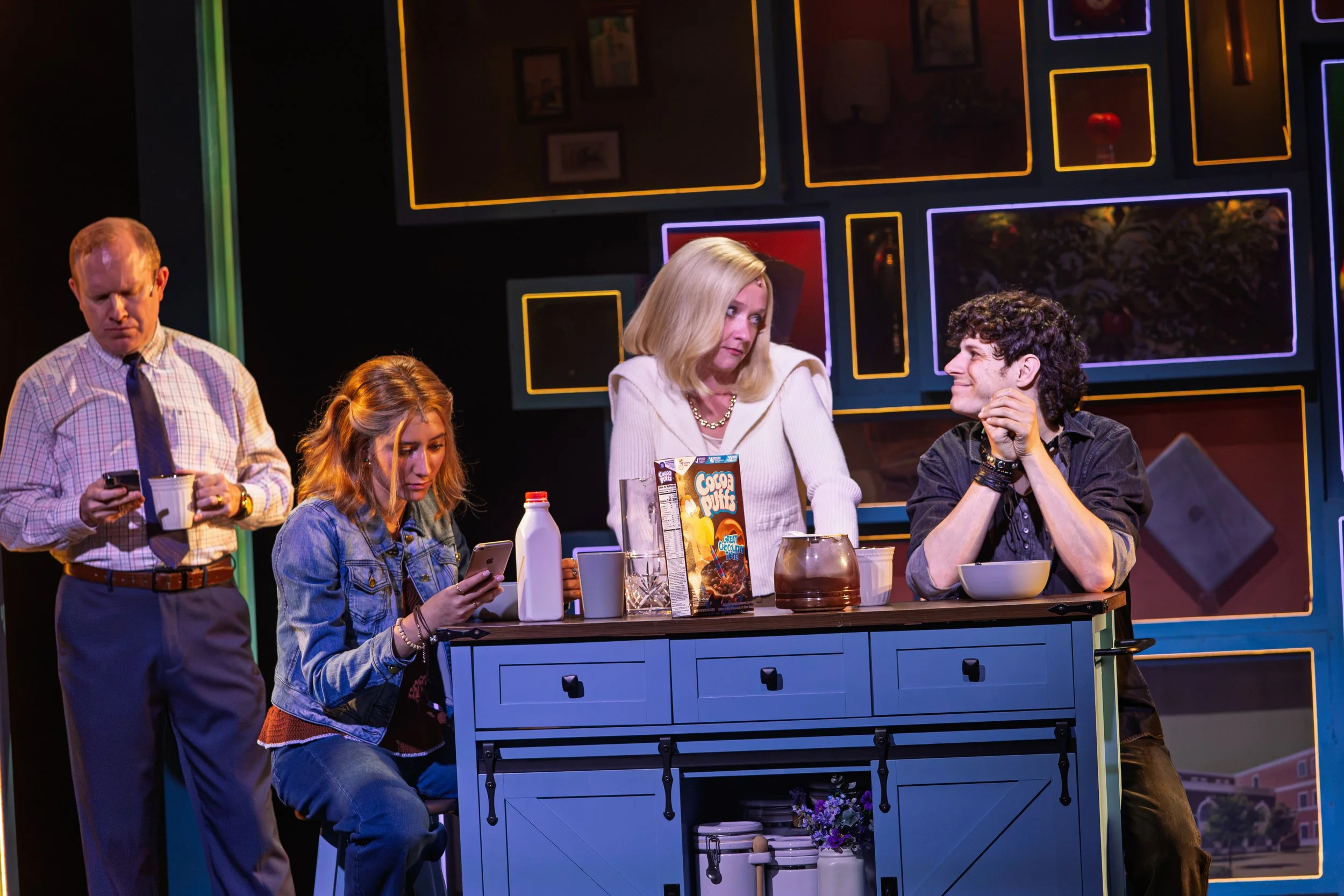 Group of five people around a kitchen island with colorful framed artwork on the wall behind them. One man is looking at his phone, a woman is looking at her phone, a woman is reaching for a cereal box. Maggie Bera playing Cynthia Murphy in Dear Evan