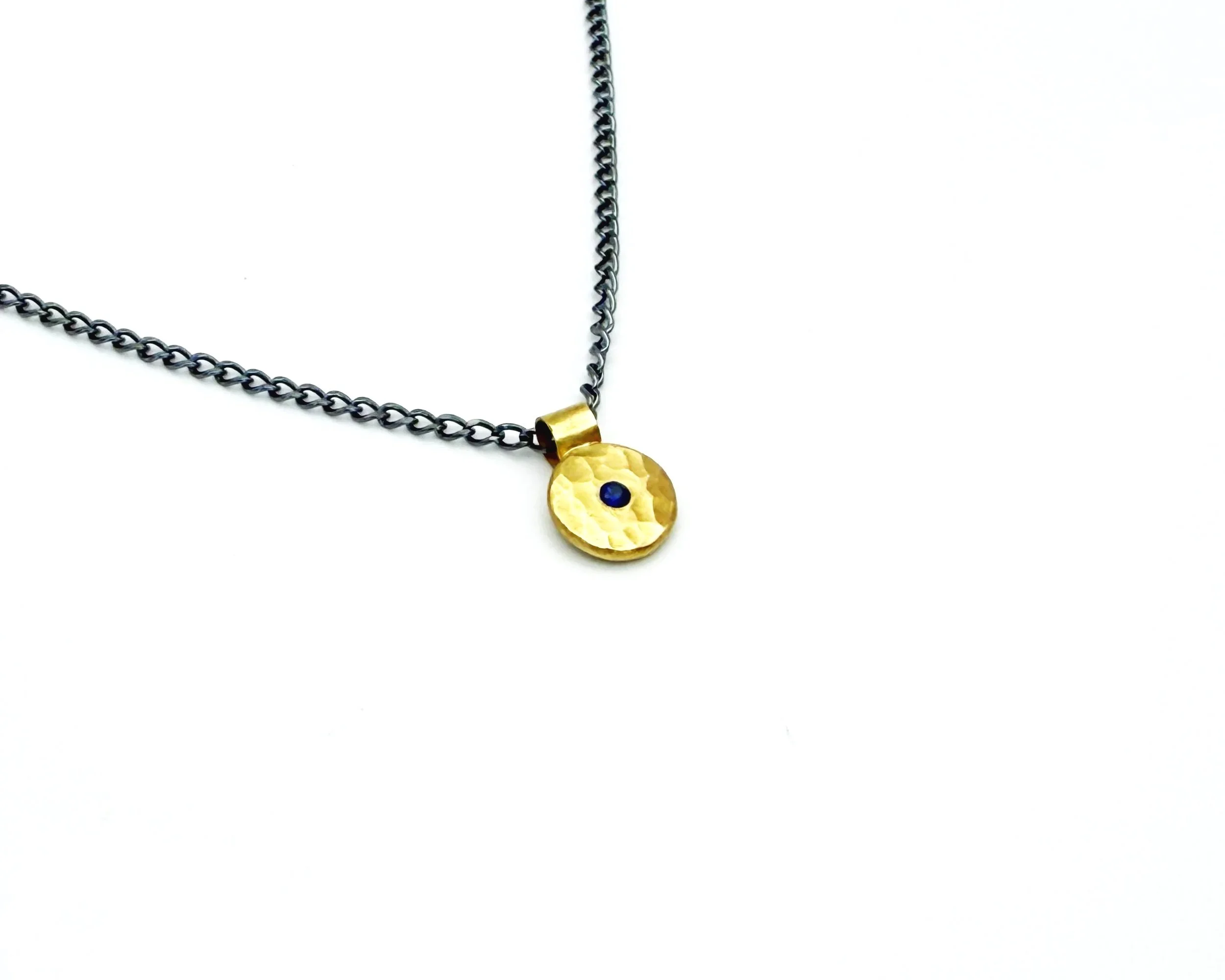 Hammered 18k gold disc pendant with a 2mm blue sapphire on an oxidized silver chain, on white