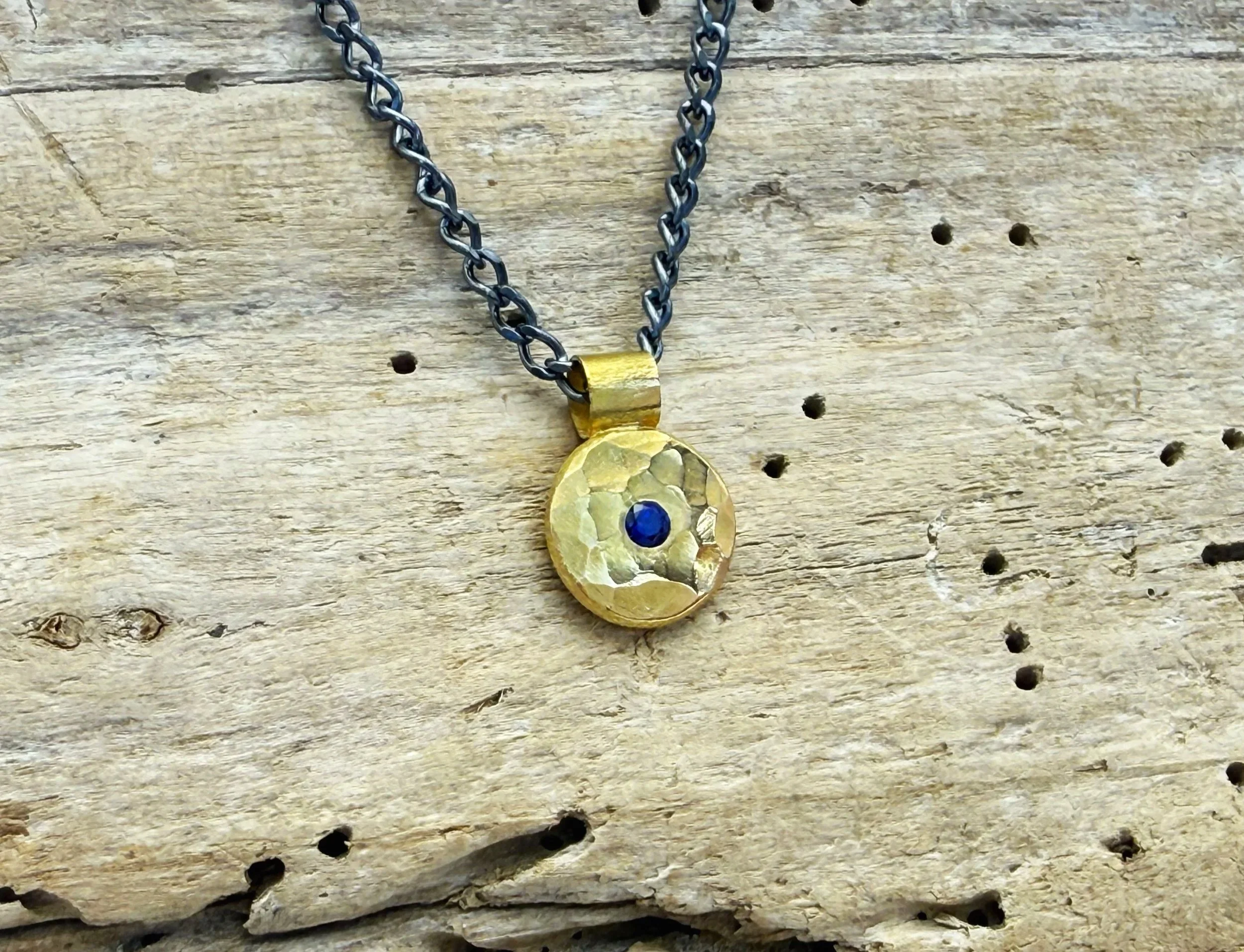 Close-up of the hammered 18k gold disc with deep blue sapphire, on weathered wood