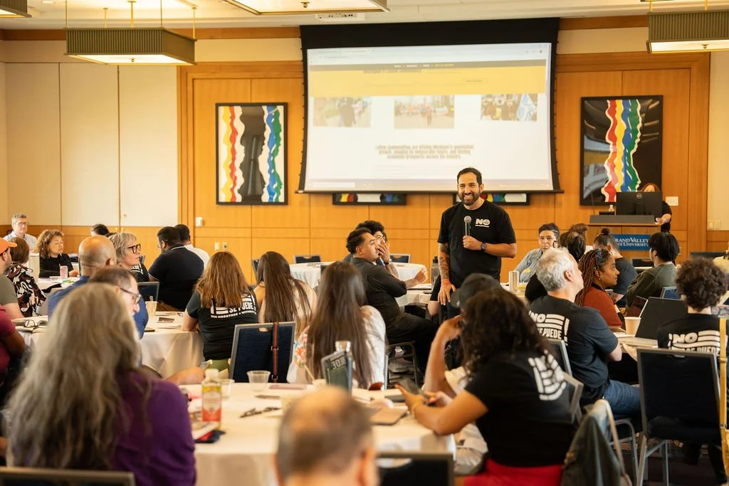 A man holding a microphone presents at a conference room filled with audience members seated at round tables, with a large screen displaying a presentation behind him.