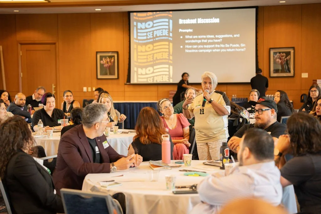 A diverse group of people seated around round tables in a conference room, attentively listening to a woman speaking with a microphone in front of a projector screen displaying a presentation titled 'Breakout Discussion'.
